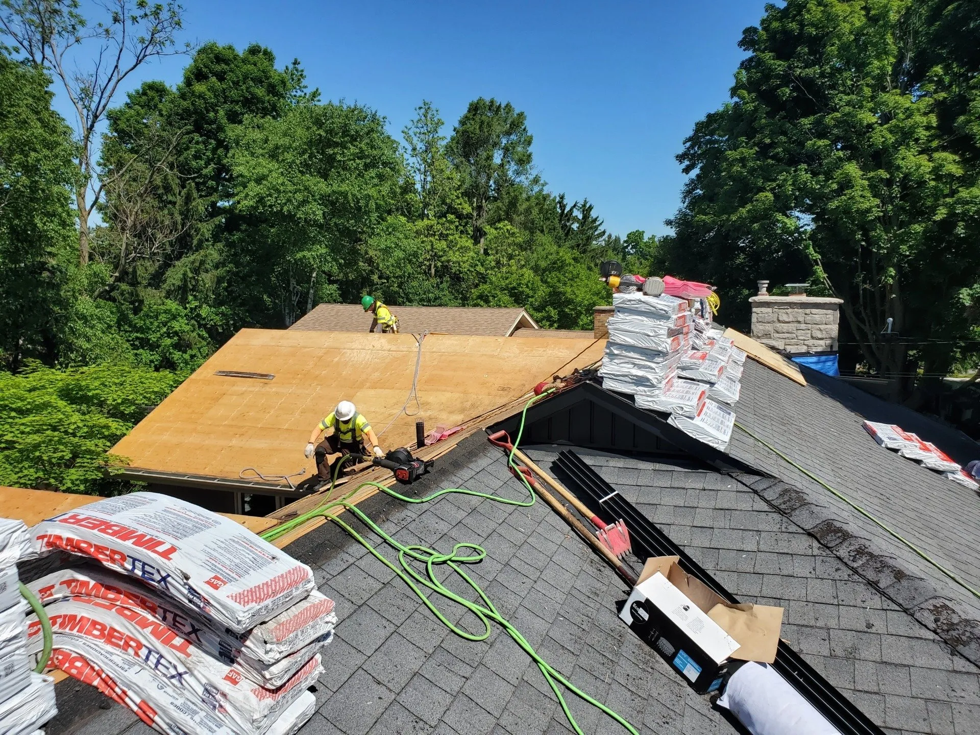 Two people are working on a half-repaired roof on a sunny day, surrounded by trees. Roofing materials and safety equipment are visible around them.