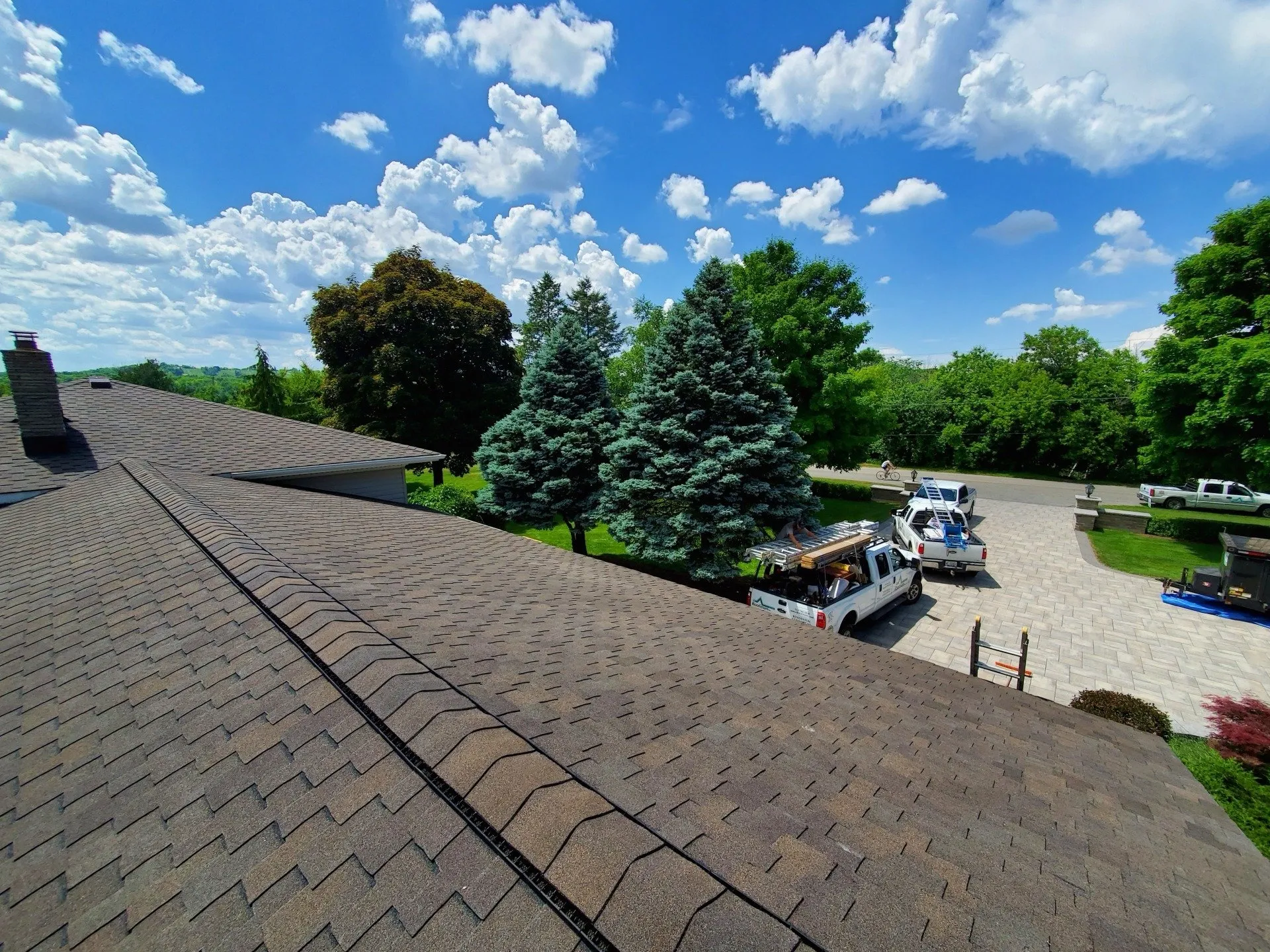 A view from a rooftop featuring shingles in the foreground, with lush green trees, utility vehicles, and a clear sky with fluffy clouds above.