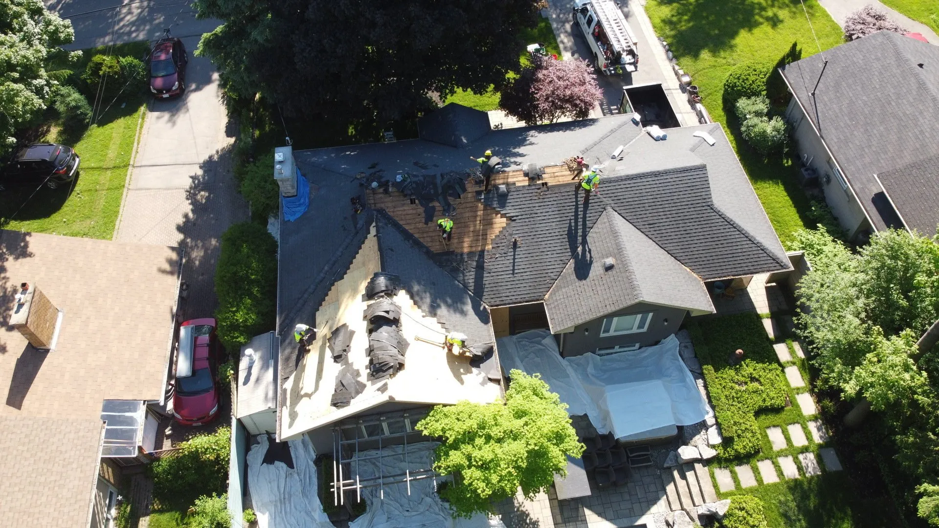 Aerial view of a suburban home where several people are working on the roof, with a mix of removed and intact shingles and construction materials around.