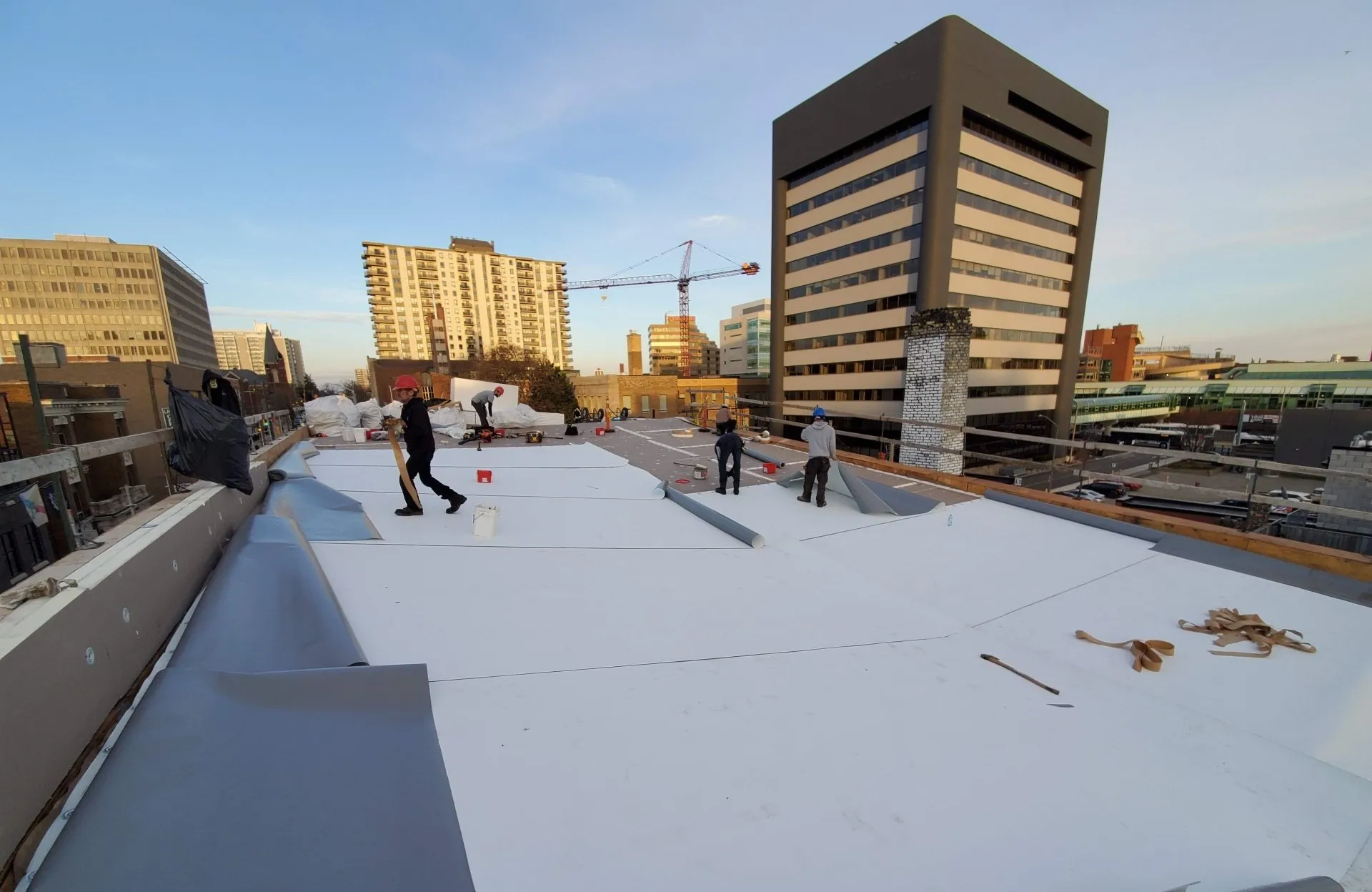 Several persons are working on a rooftop at dusk, with city buildings and a construction crane in the background. The scene depicts an urban landscape.