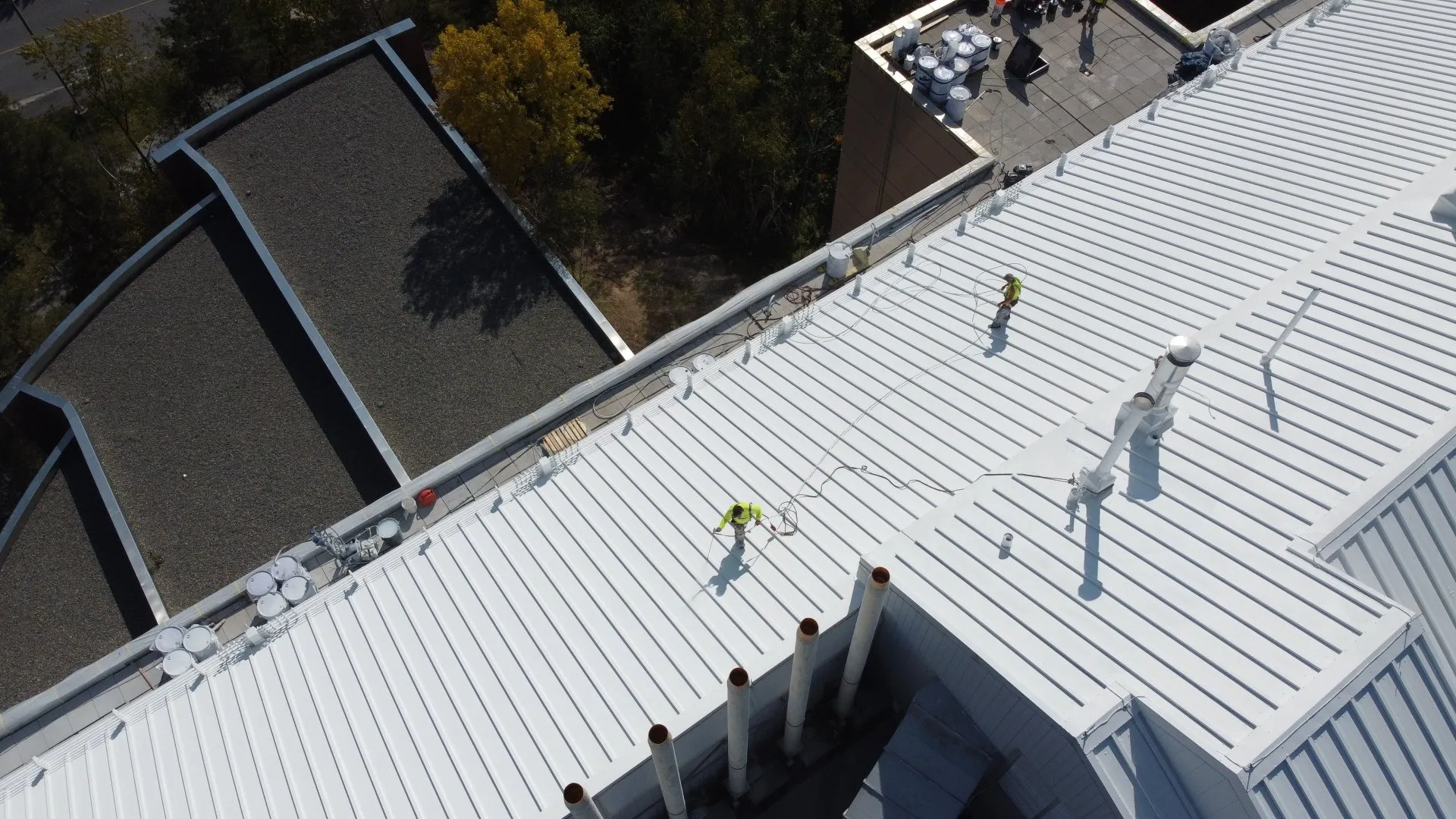 Aerial view of two people in safety harnesses working on a corrugated metal rooftop, with ventilation pipes and surrounding greenery visible.