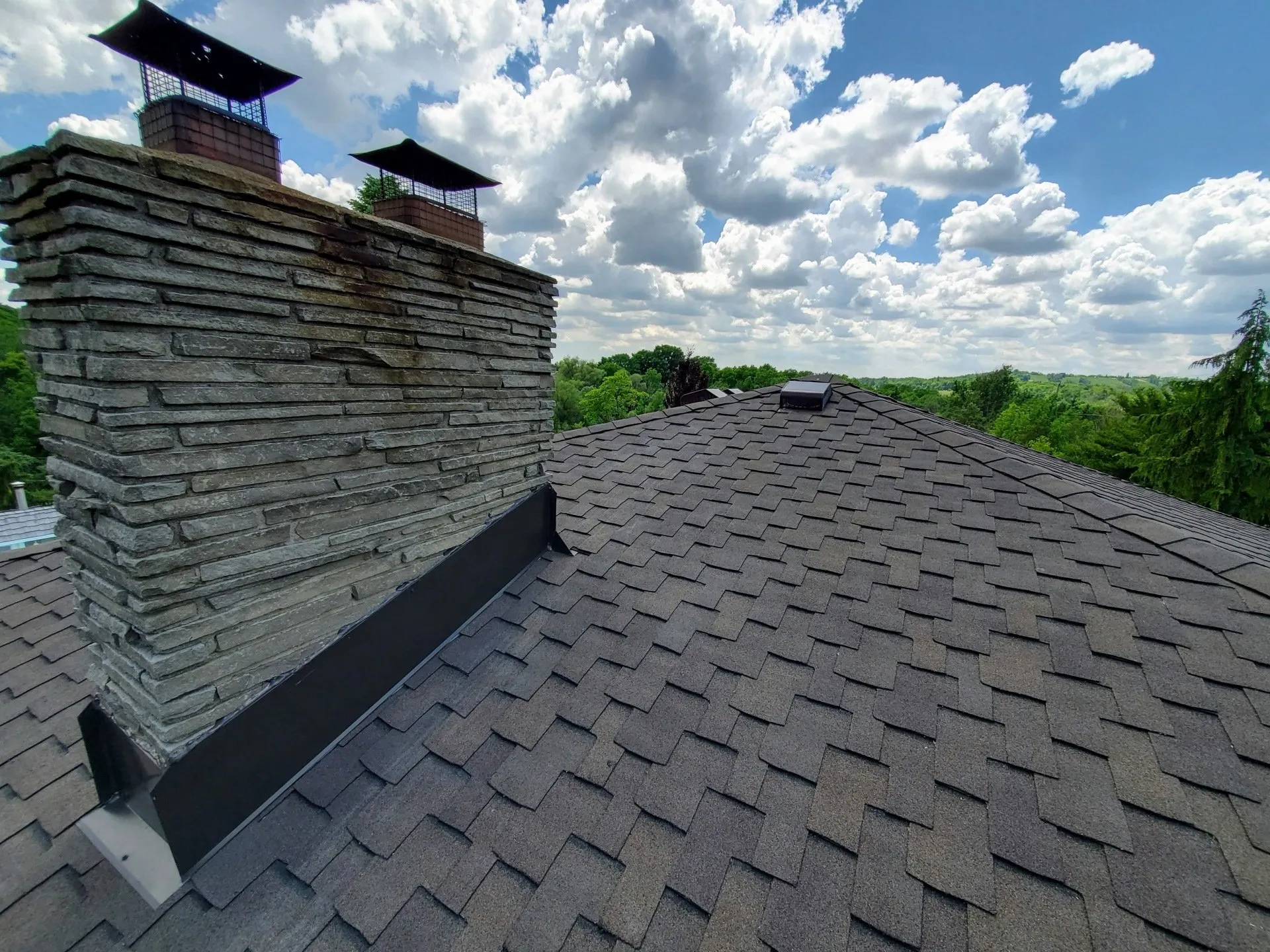 An expansive roof under a blue sky with clouds features a large stone chimney, lush trees in the distance, and a protective chimney guard.