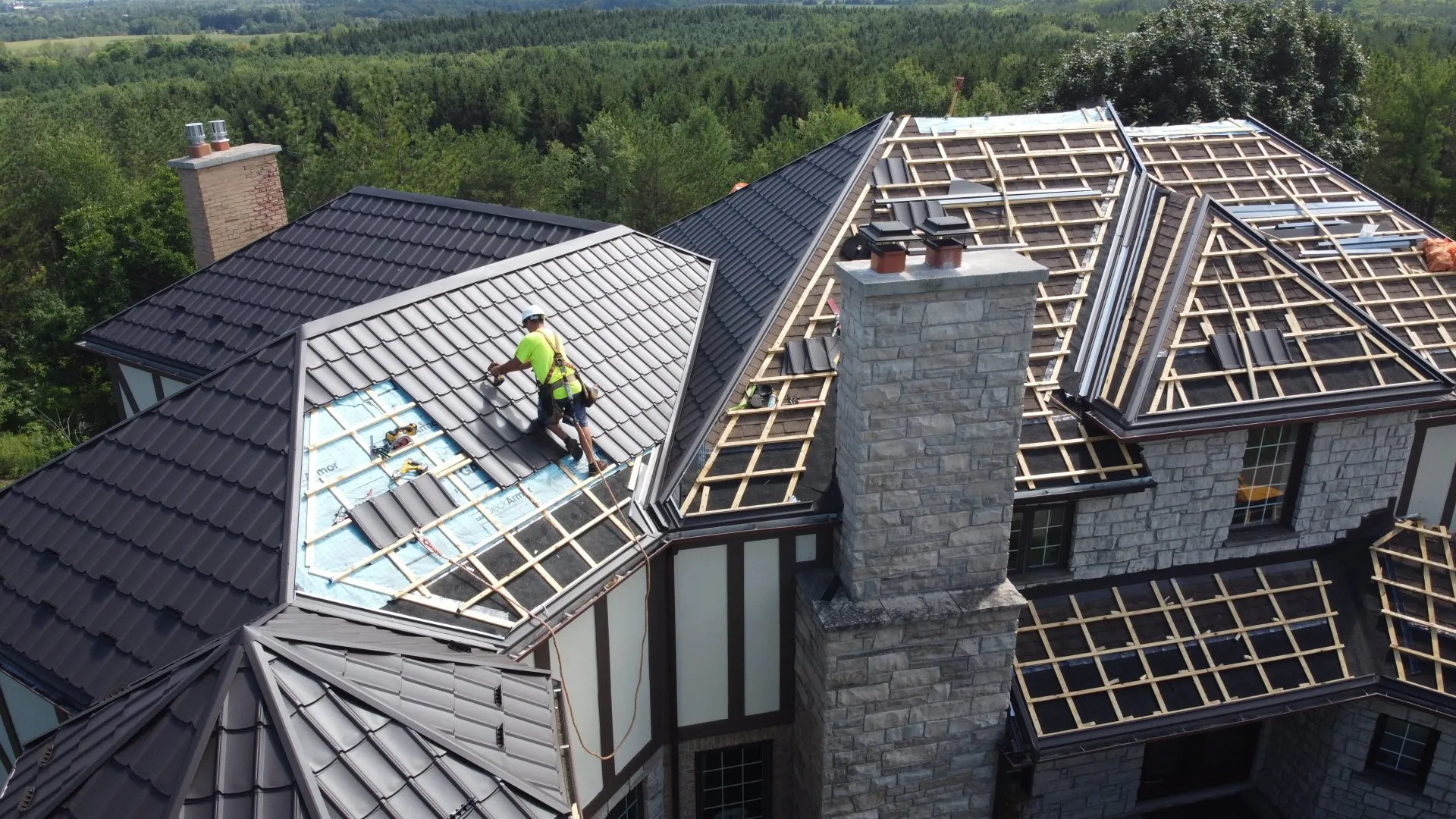 A person is working on a complex, multi-level roof with partially installed dark tiles and exposed wooden beams, set amidst a lush green forest.