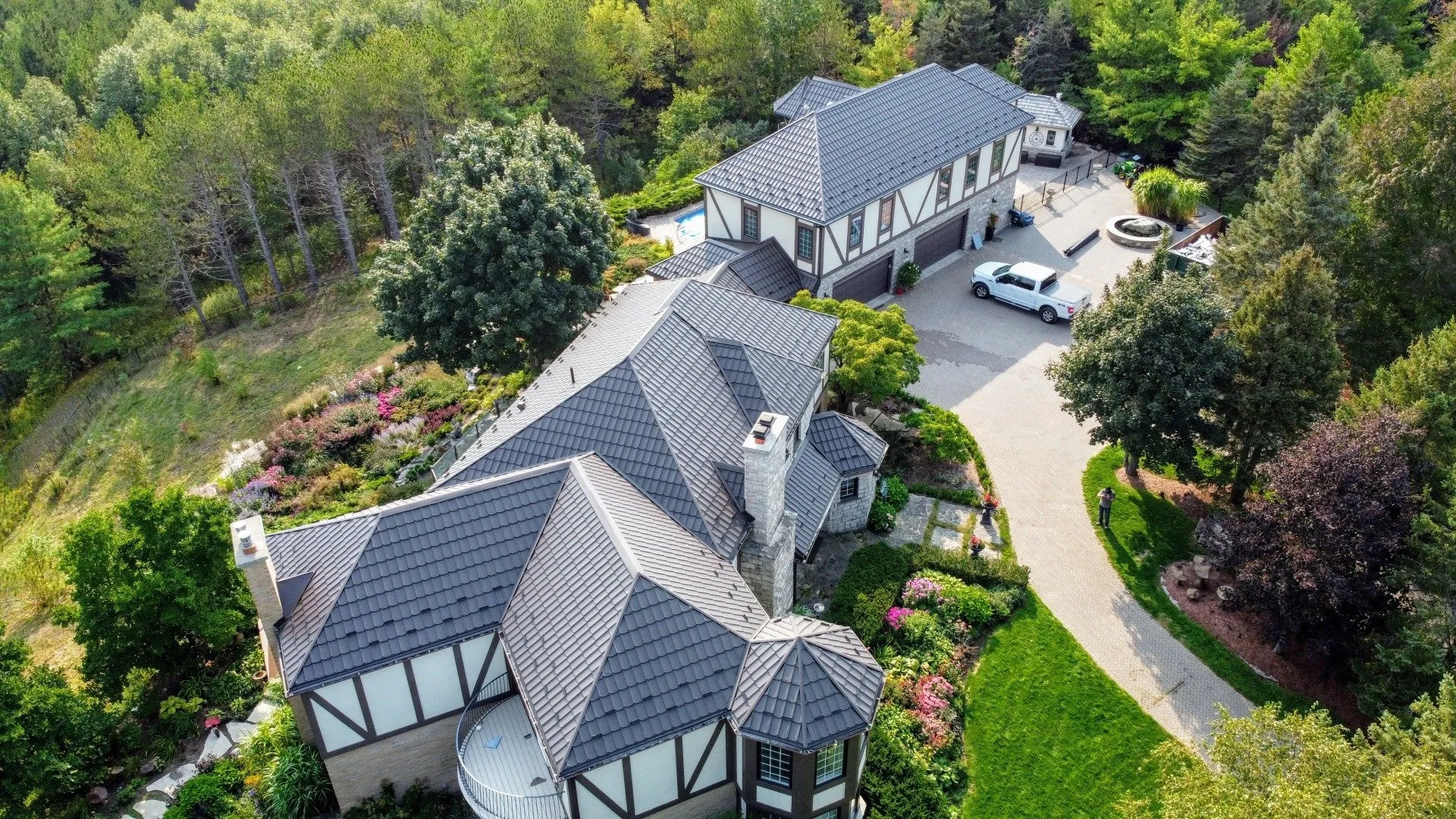 Aerial view of a large, multi-level house with grey shingles, surrounded by lush greenery, landscaped garden, driveway, and a white vehicle parked outside.