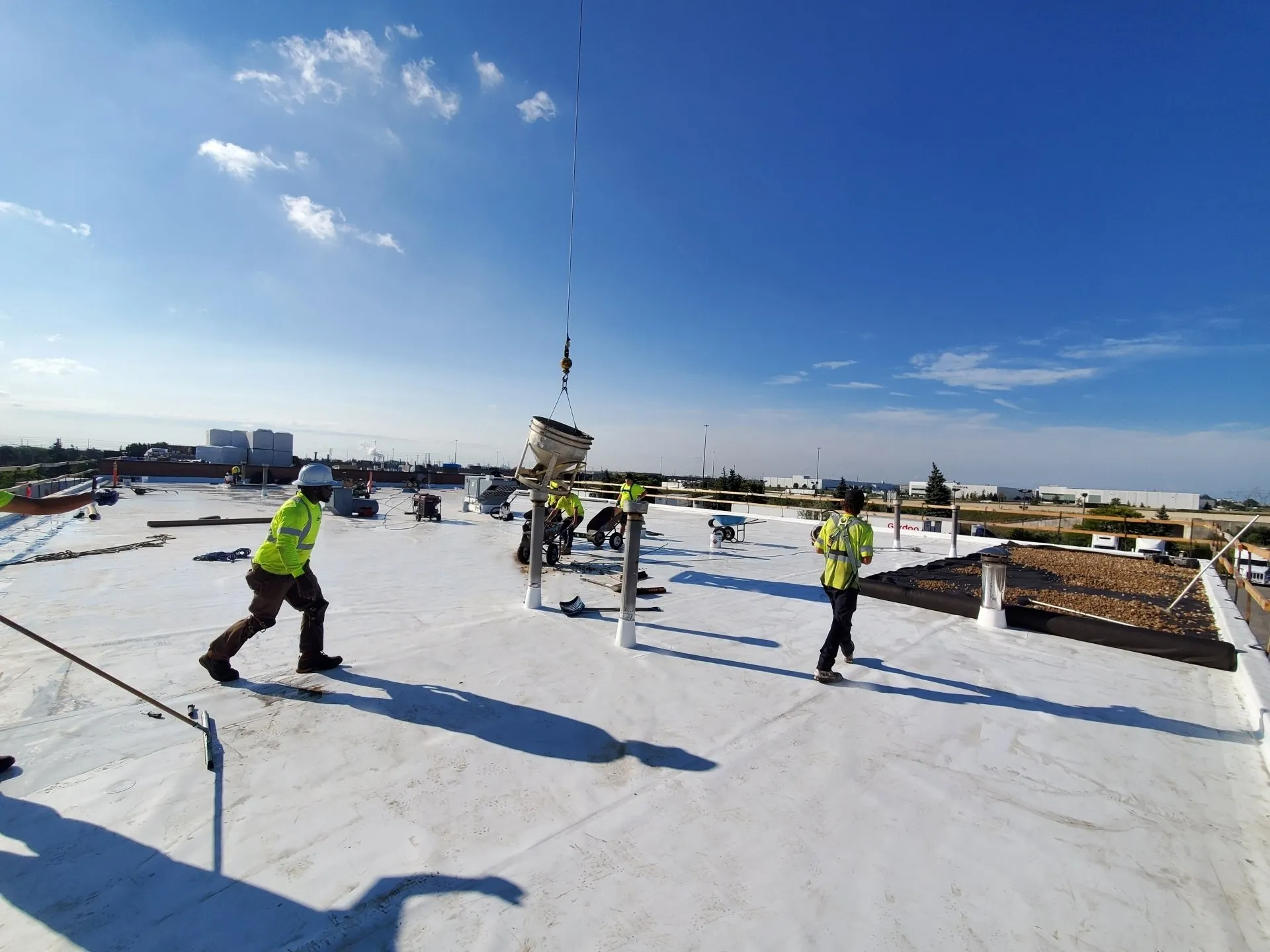 This image shows multiple people in high-visibility jackets working on a rooftop construction site under a clear blue sky with materials being hoisted.