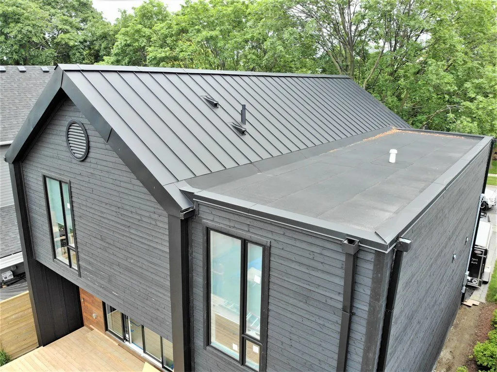 Aerial view of a modern house with a dark metal roof and gray siding. The structure features large windows, a flat section, and greenery around.