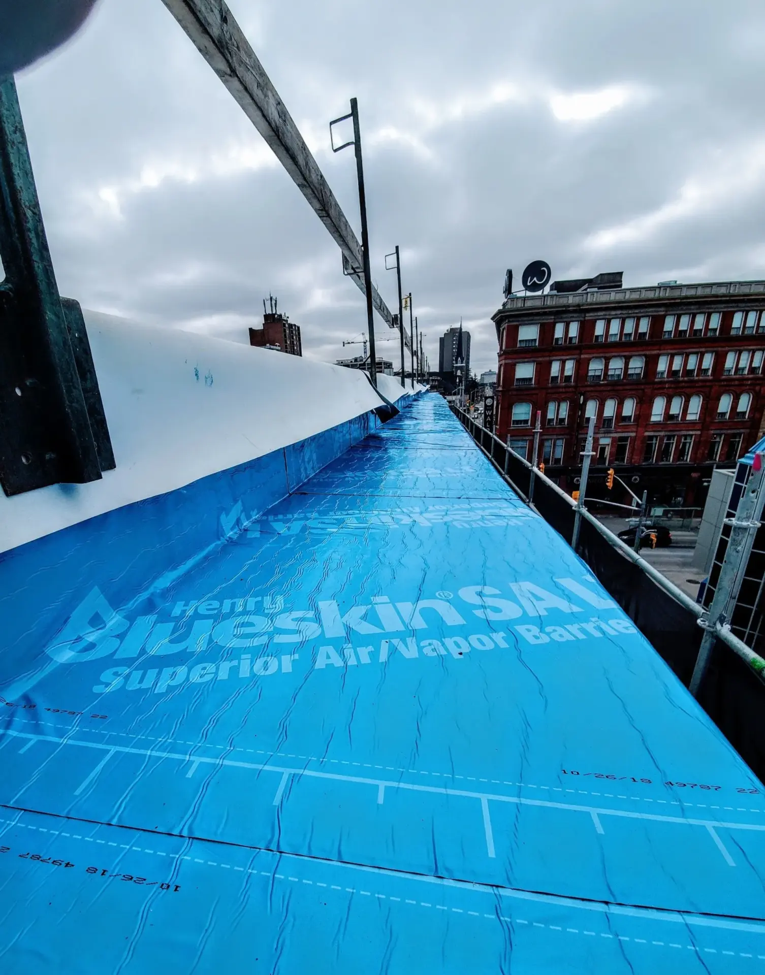 View from above on a construction site with blue protective covering and urban buildings in the background under an overcast sky.