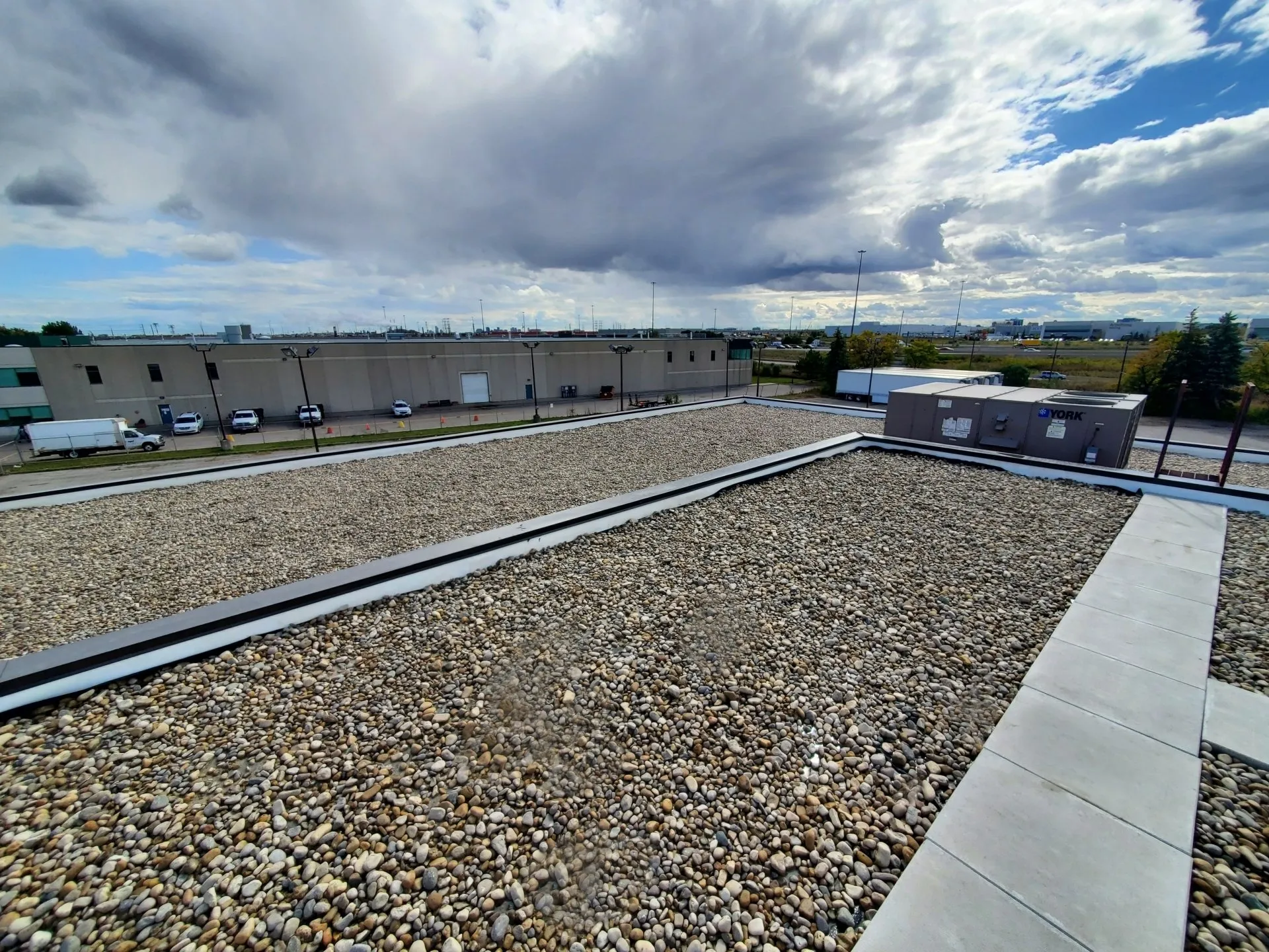 The image shows an industrial area as viewed from a rooftop covered with gravel. Warehouses, parked trucks, and cloudy skies are visible in the background.