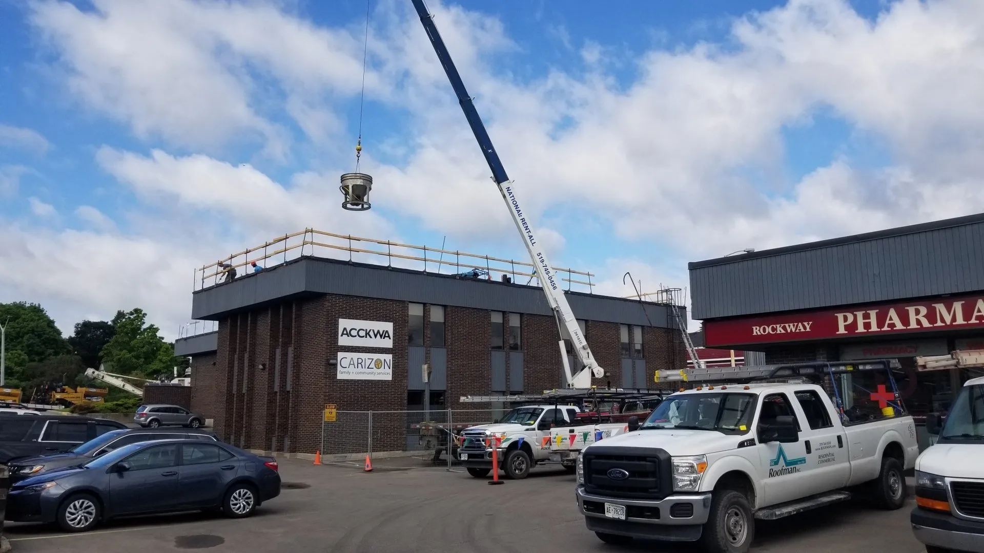 A crane lifts a large object onto a building's rooftop while multiple vehicles occupy the parking lot below under a partly cloudy sky.