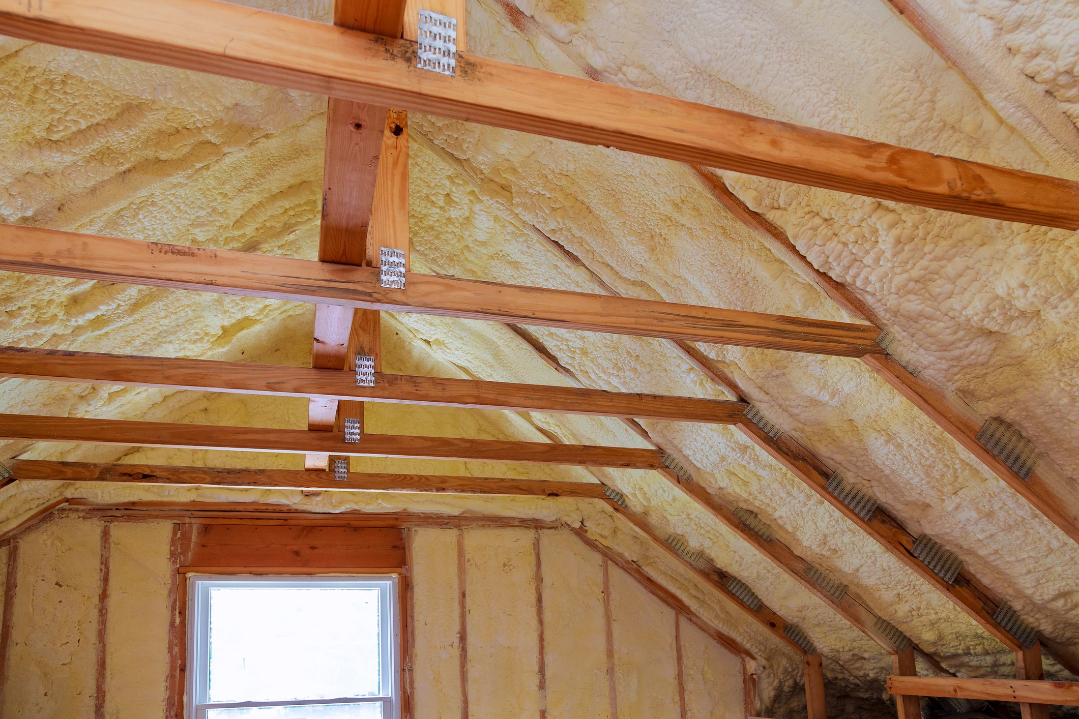 This image shows a partially finished attic with exposed wooden beams and yellow insulation foam sprayed on the underside of the roof.