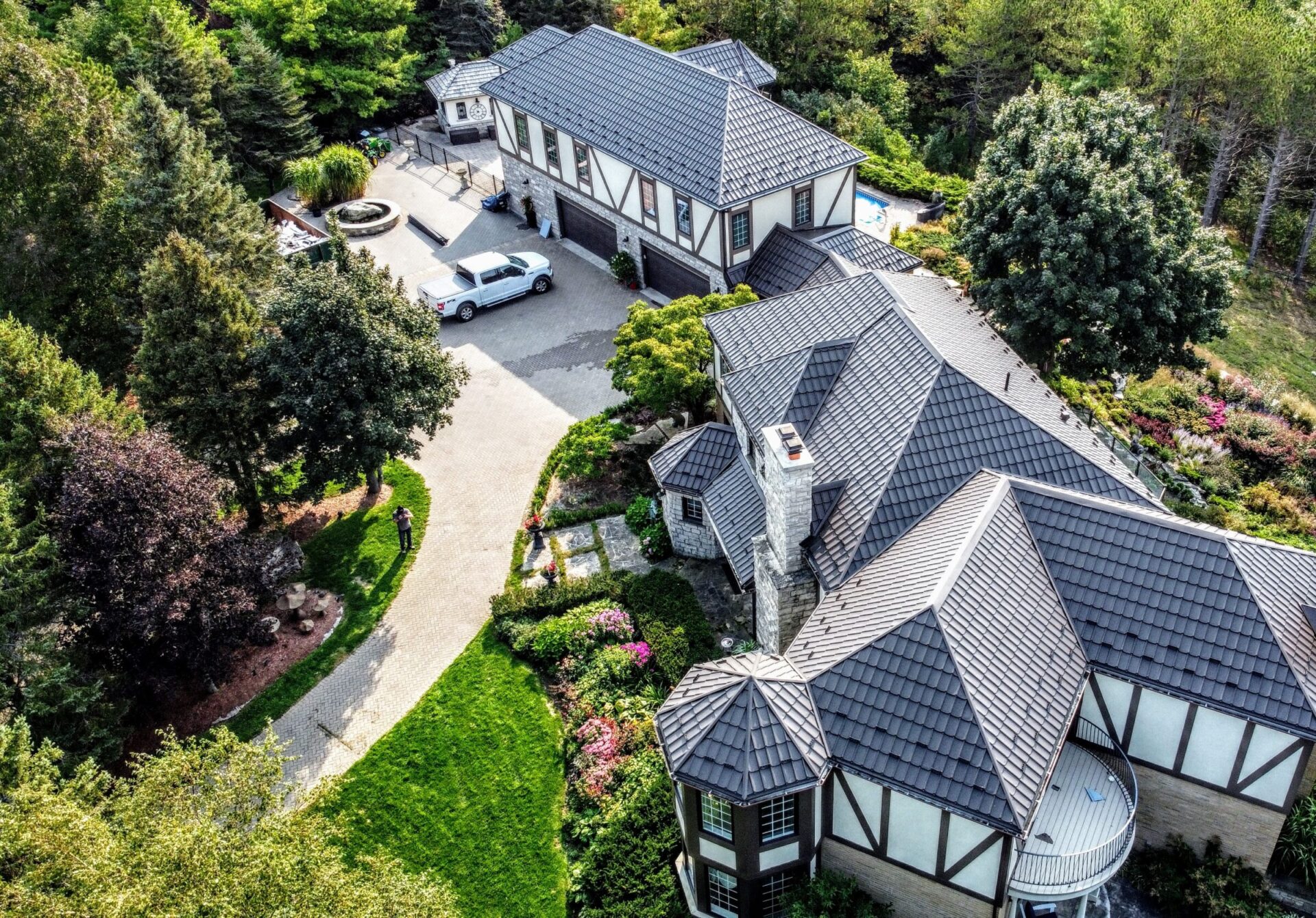 Aerial view of a luxurious house with a steeply pitched roof, surrounded by manicured gardens, trees, and a driveway with a parked vehicle.