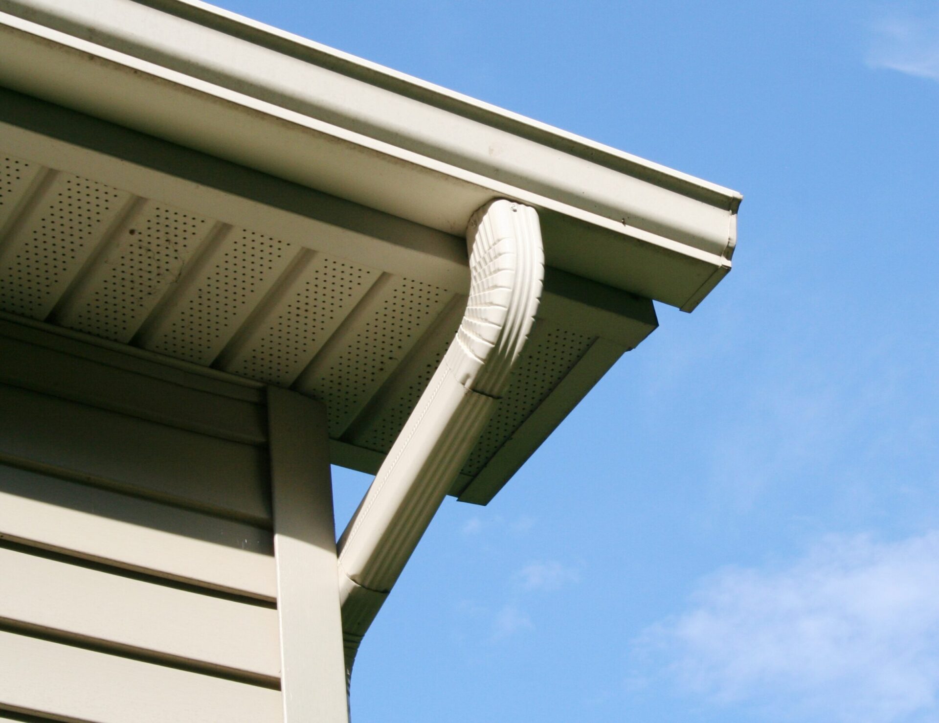 A beige house's soffit and gutter under a clear blue sky. The downspout connects awkwardly to the gutter, suggesting improper installation or damage.