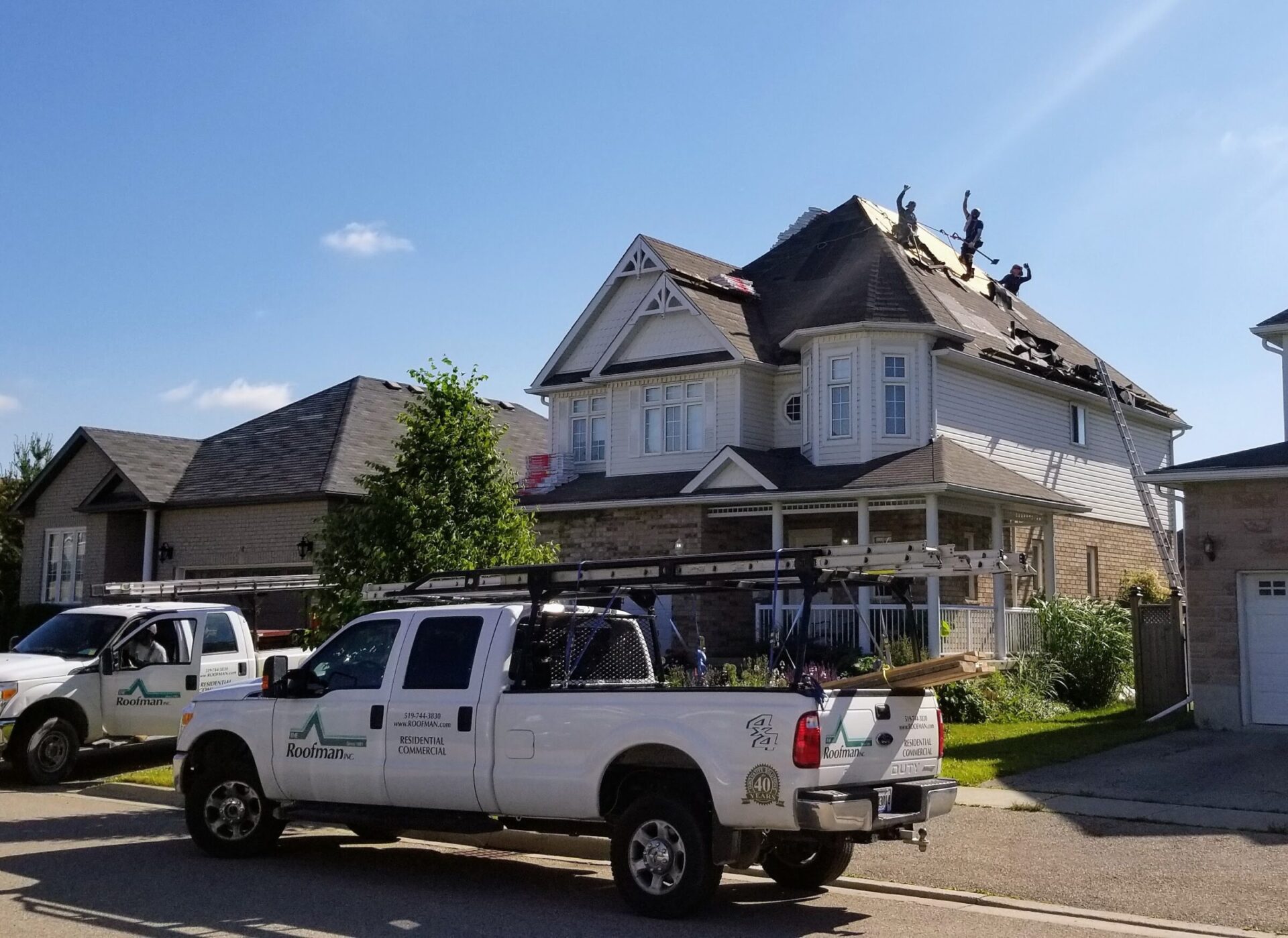 Several people working on a residential roof, with a roofing company's truck parked below. The roof is partly stripped, under a clear blue sky.