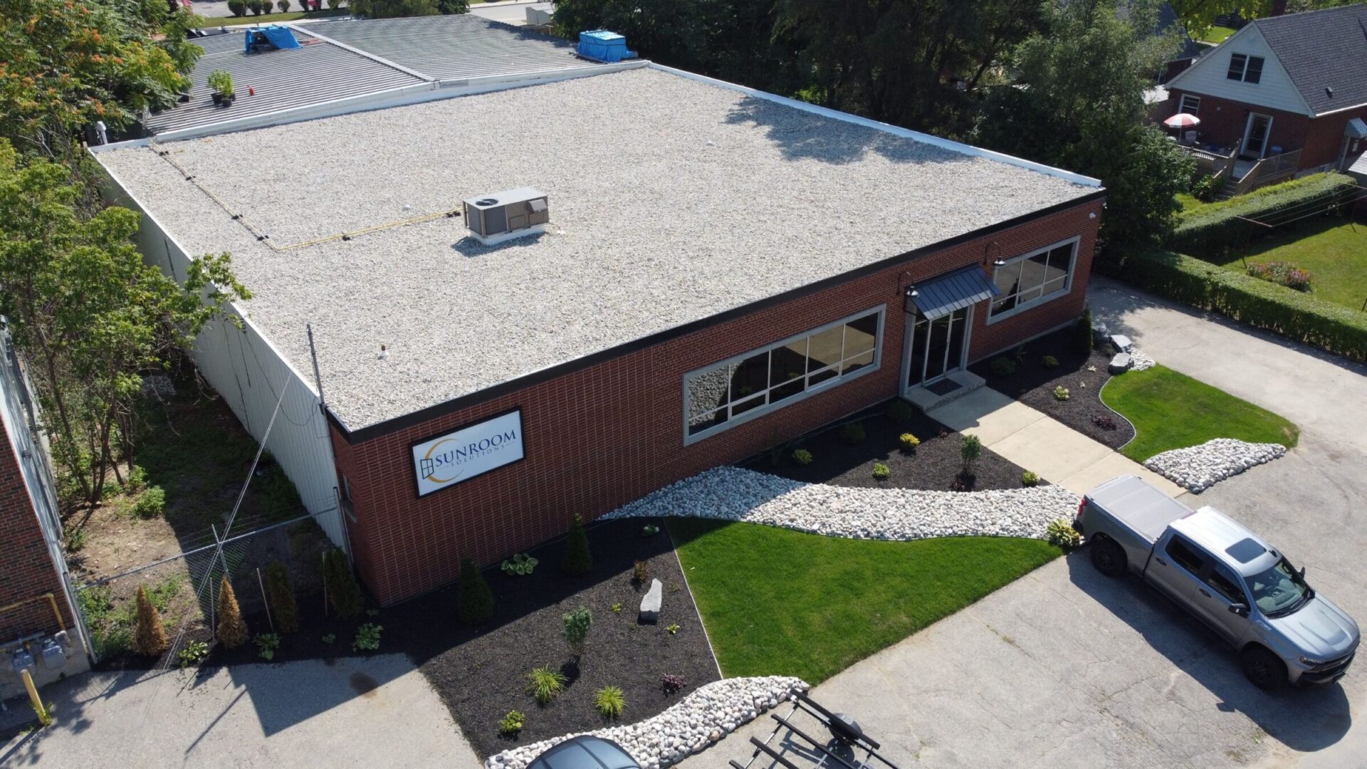 Aerial view of a single-story brick building with a gravel-covered flat roof, landscaped with green lawn and white stones, and a parked vehicle.