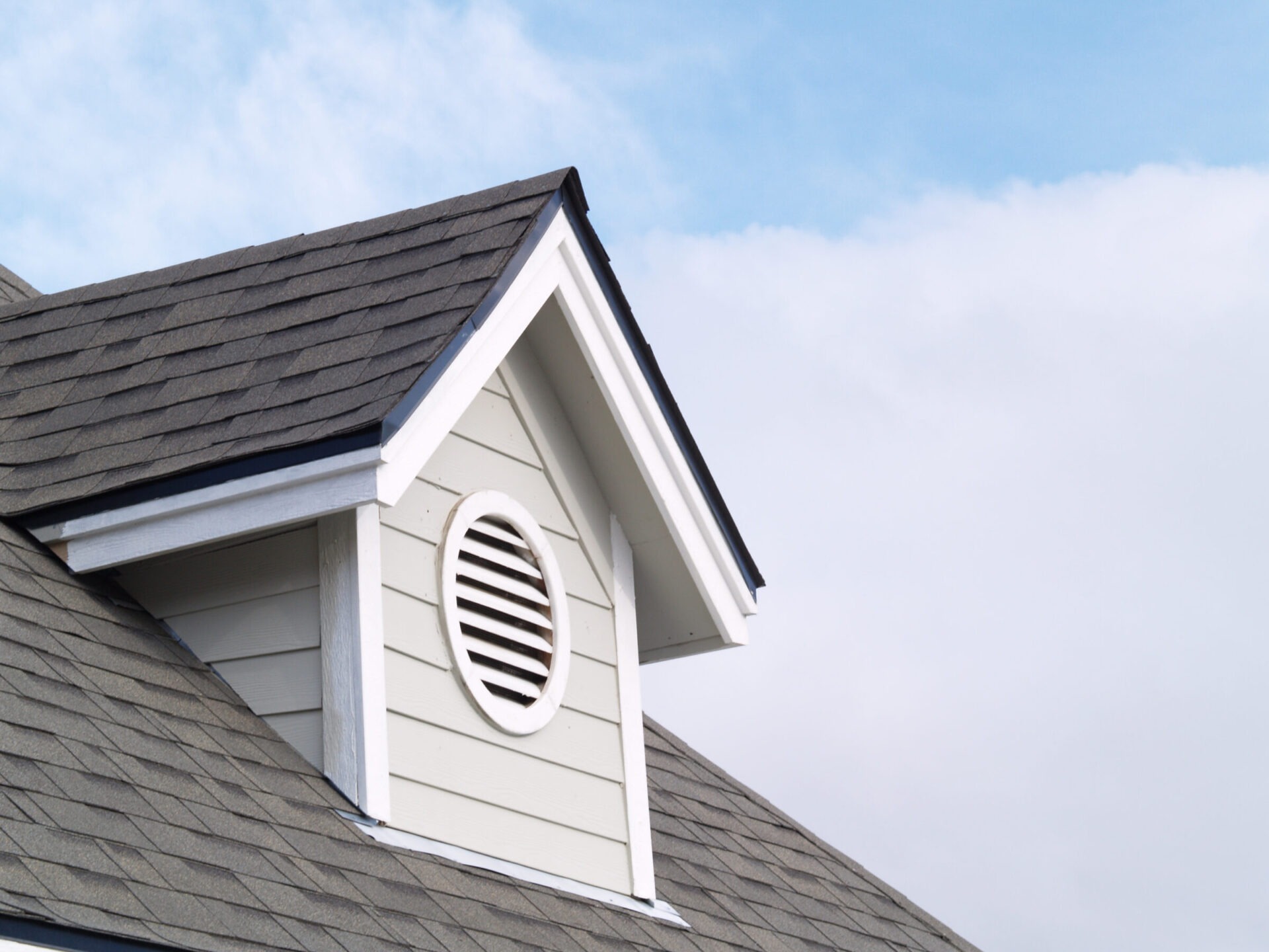 This image shows the upper portion of a house with an asphalt shingle roof, white siding, a vent, and a clear blue sky in the background.