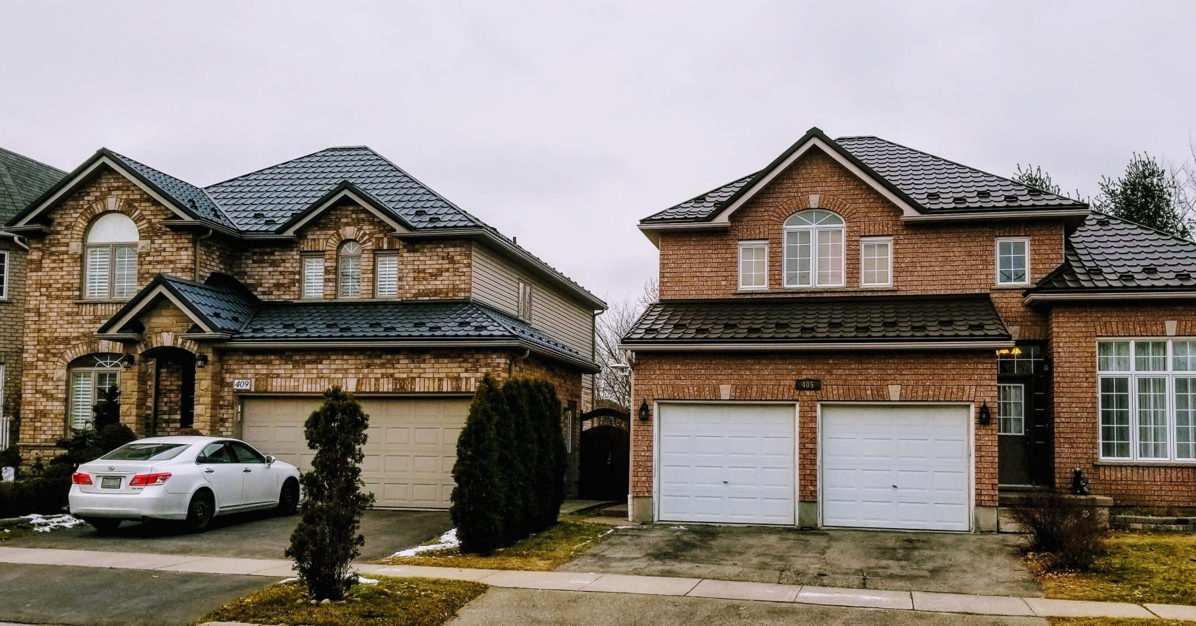 Residential street featuring two-story brick houses with multi-gabled roofs, garages, and a white sedan parked in a driveway under overcast skies.