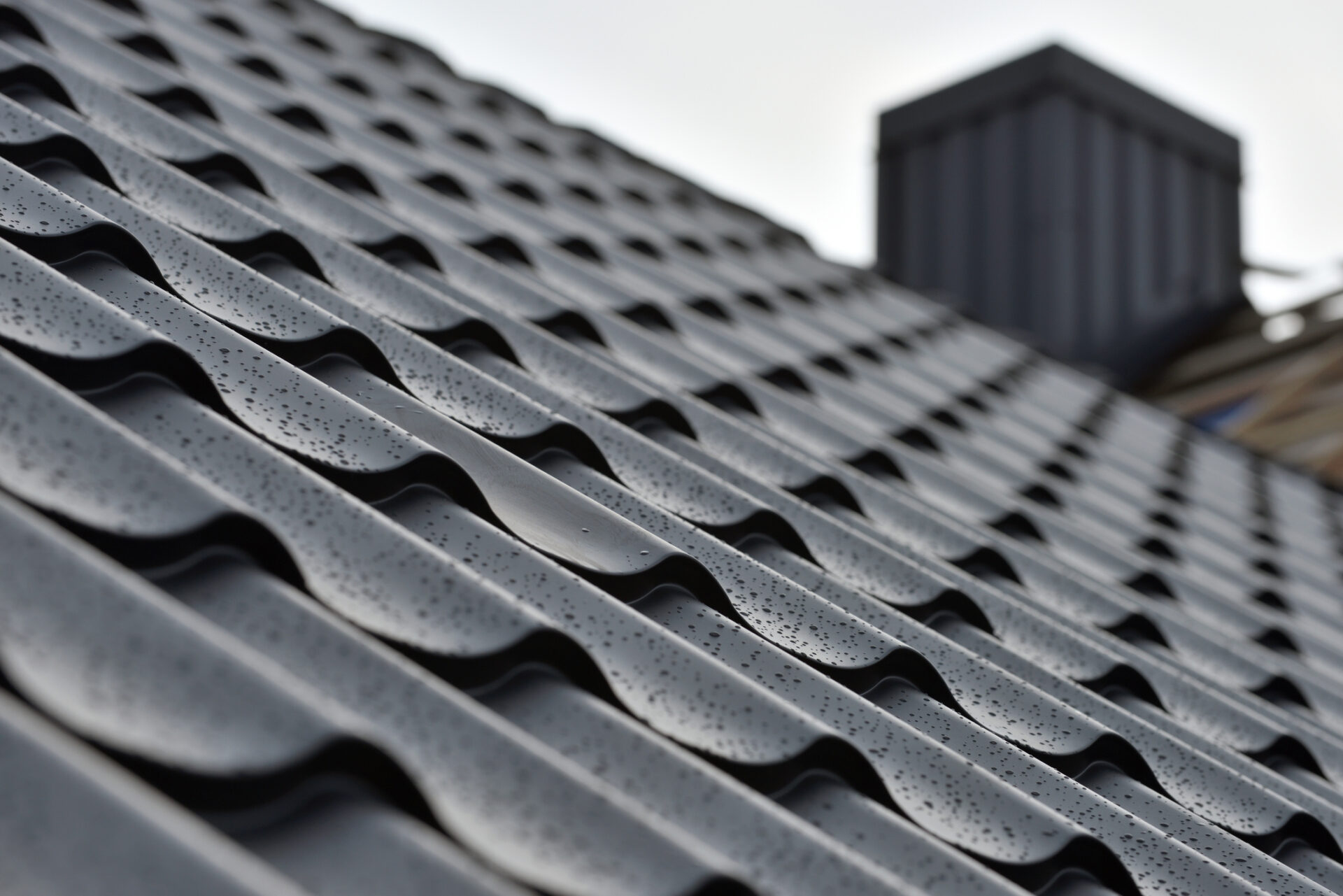 Close-up of a gray tiled roof with visible water droplets, angular perspective, beyond the rooftop there's a blurred building against a cloudy sky.