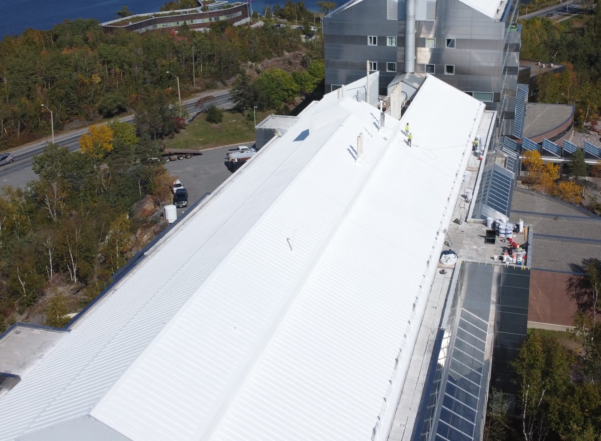 Aerial view of a large industrial building with a corrugated metal roof. People are working on the rooftop, near machinery and safety equipment. Trees and a road are visible.