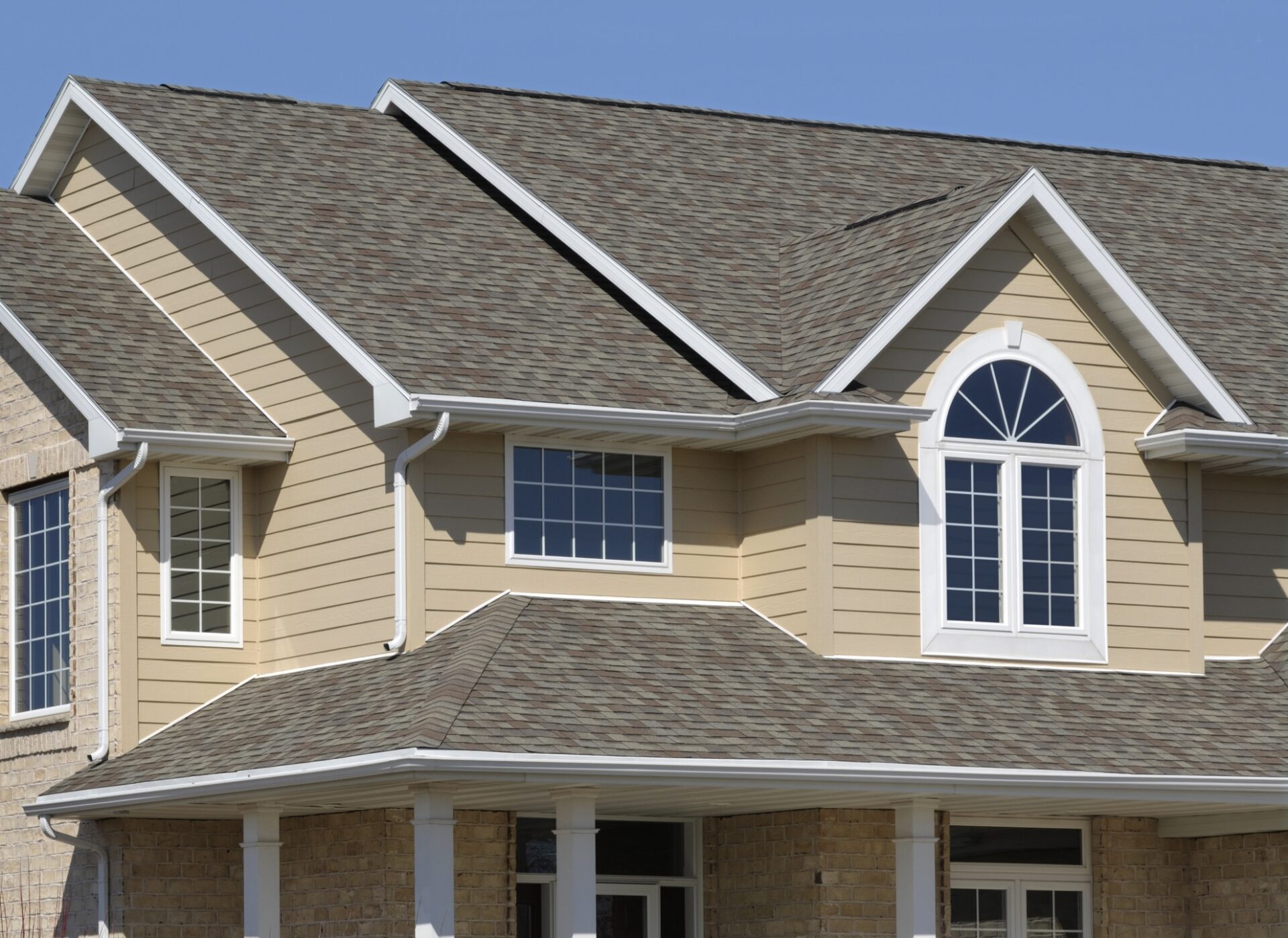 This image shows the upper facade of a suburban house with beige siding, a brown shingled roof, white trim, and several symmetric windows.