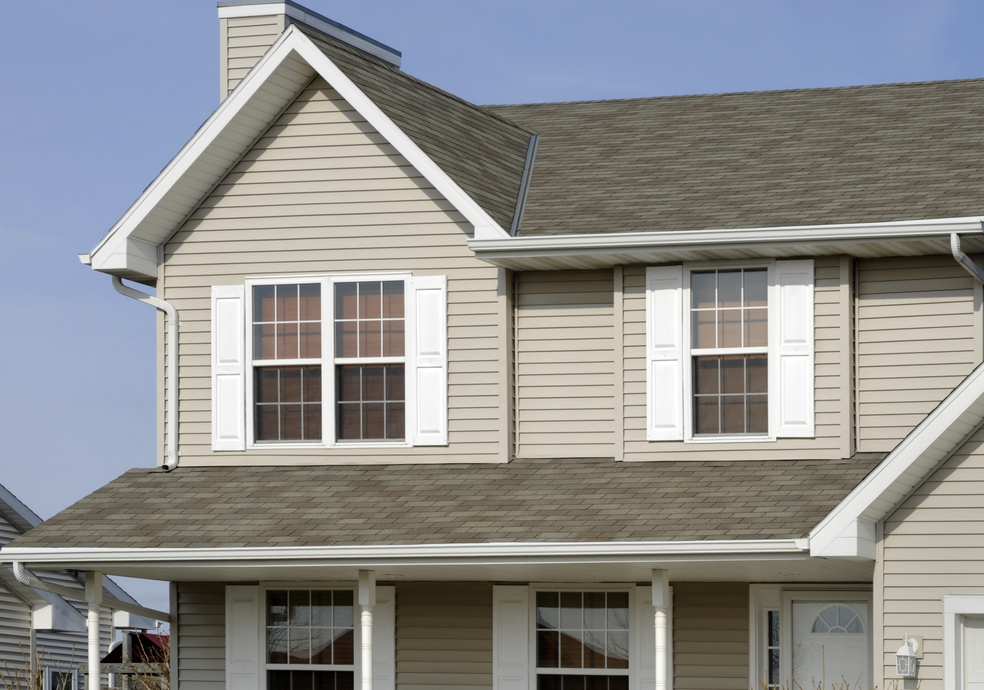 This is a two-story residential house with beige siding, white trim around windows, a shingled roof, and a clear blue sky in the background.