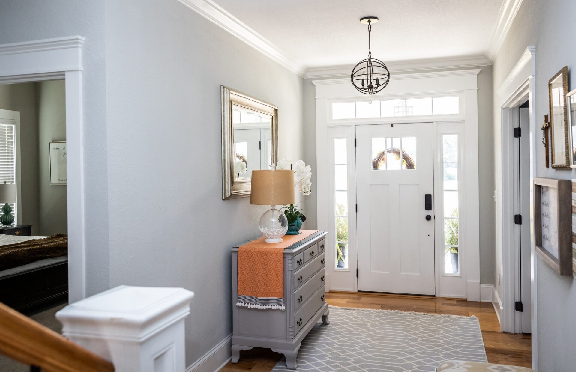 An elegant entryway with a white front door, hardwood floors, a geometric rug, a gray chest of drawers, a lamp, a mirror, and a chandelier.