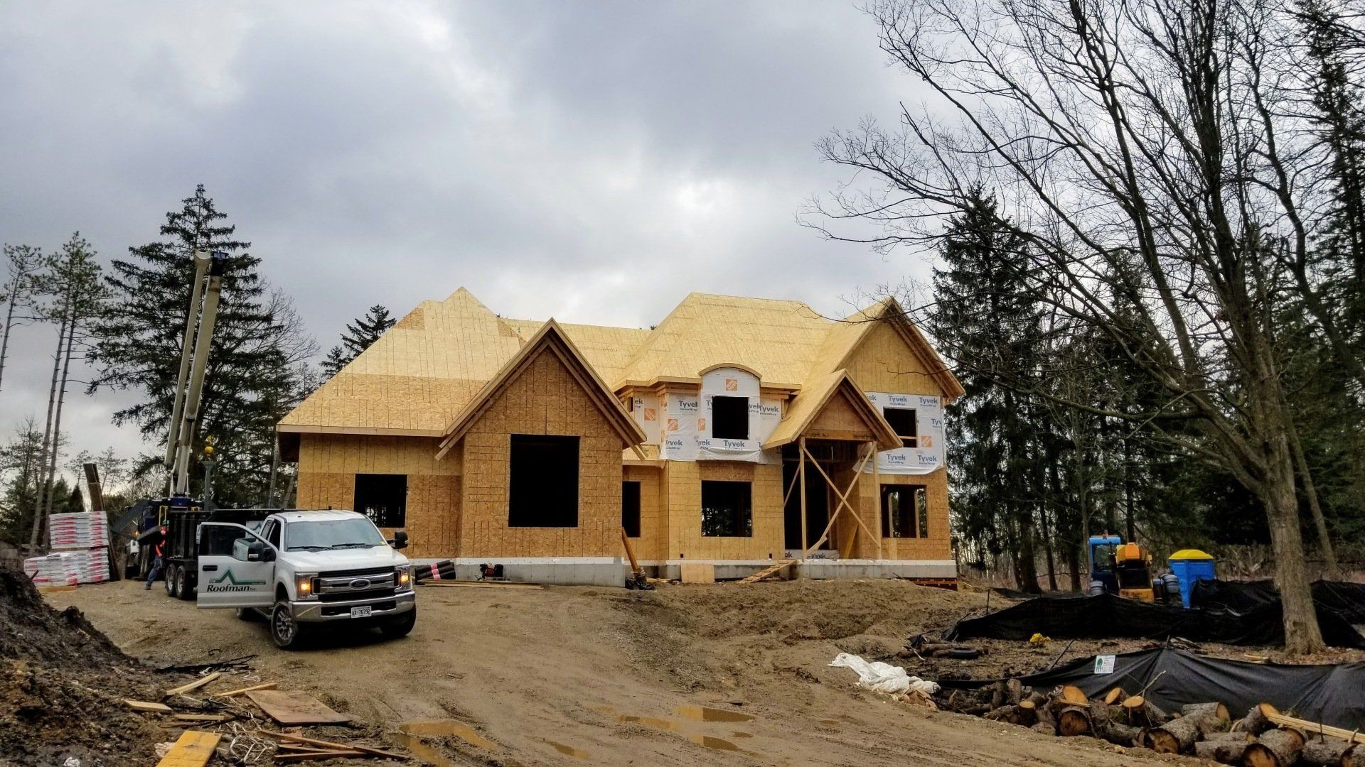 A partially constructed house with exposed plywood sheathing, surrounded by construction equipment, materials, and a pickup truck on a cloudy day.