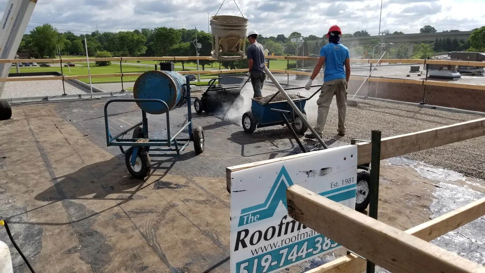 Two persons are performing roofing work; one is using a wheelbarrow while the other operates machinery, lifting a bucket. Signage indicates a roofing company.