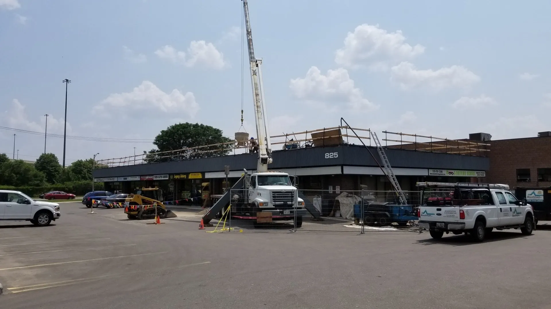 Construction site at a commercial building, with a crane lifting materials, workers on the roof, vehicles, and safety fences in a parking lot.