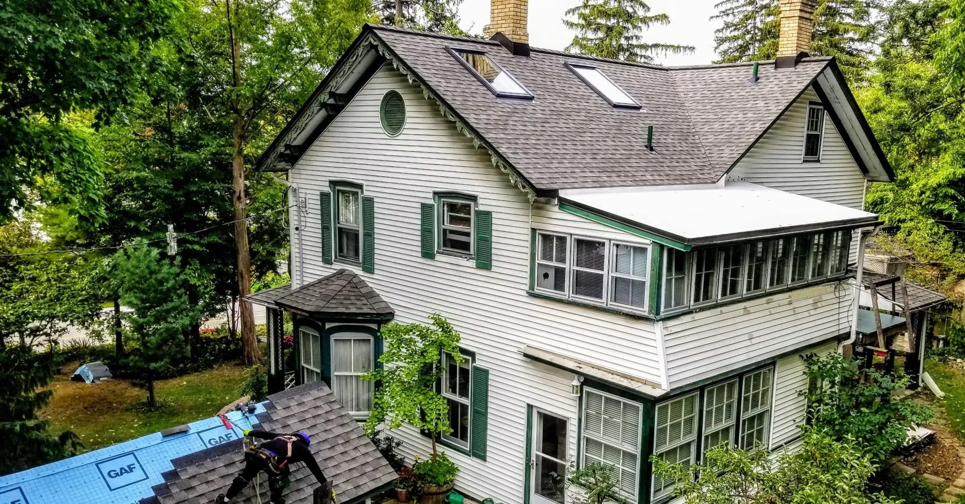 A two-story house with white siding and green shutters, surrounded by trees, with roof work in progress under a clear sky.