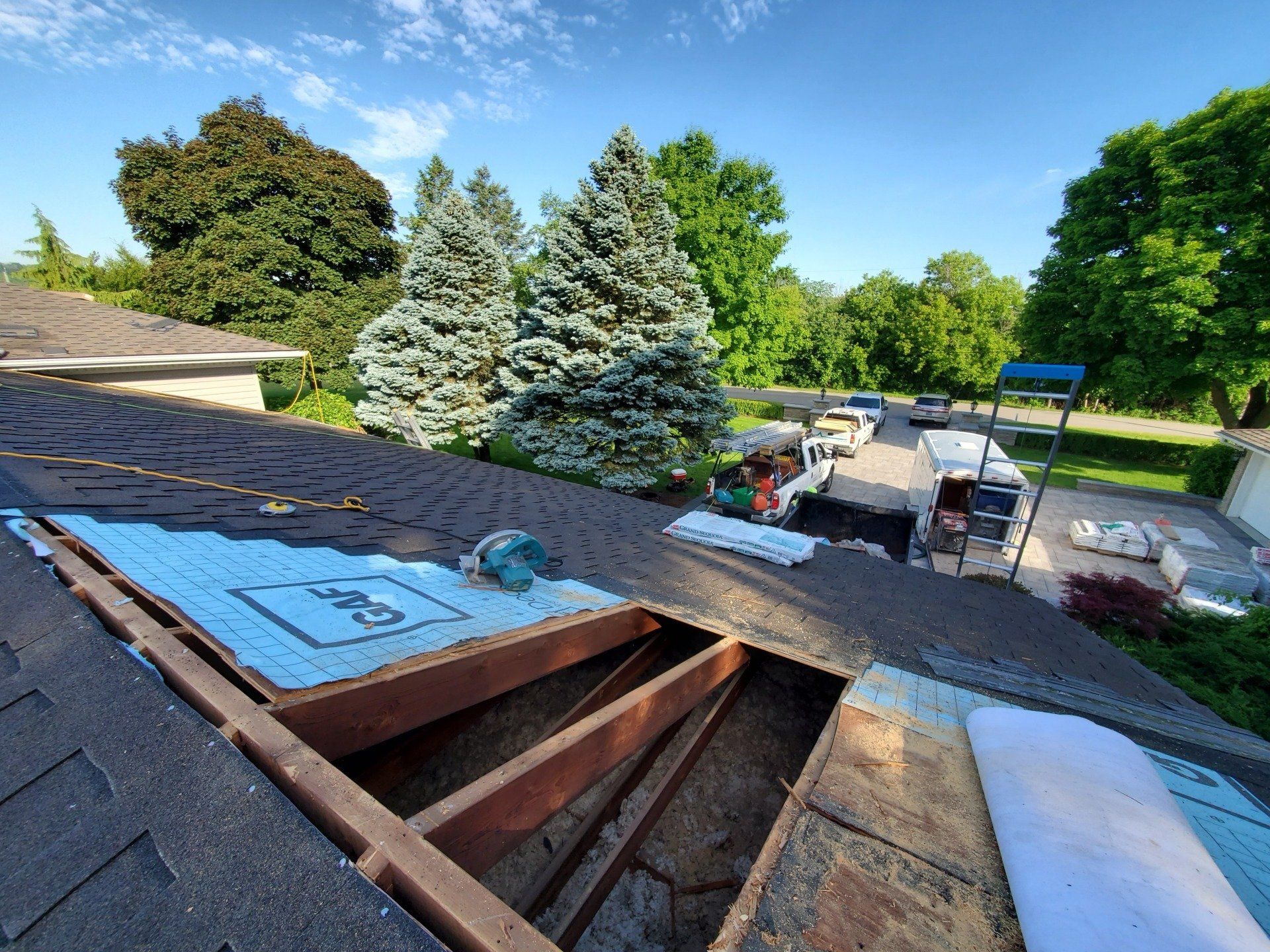 A partial roof reconstruction scene showing exposed wooden beams, construction materials, a circular saw, and vehicles parked below near a driveway with greenery.