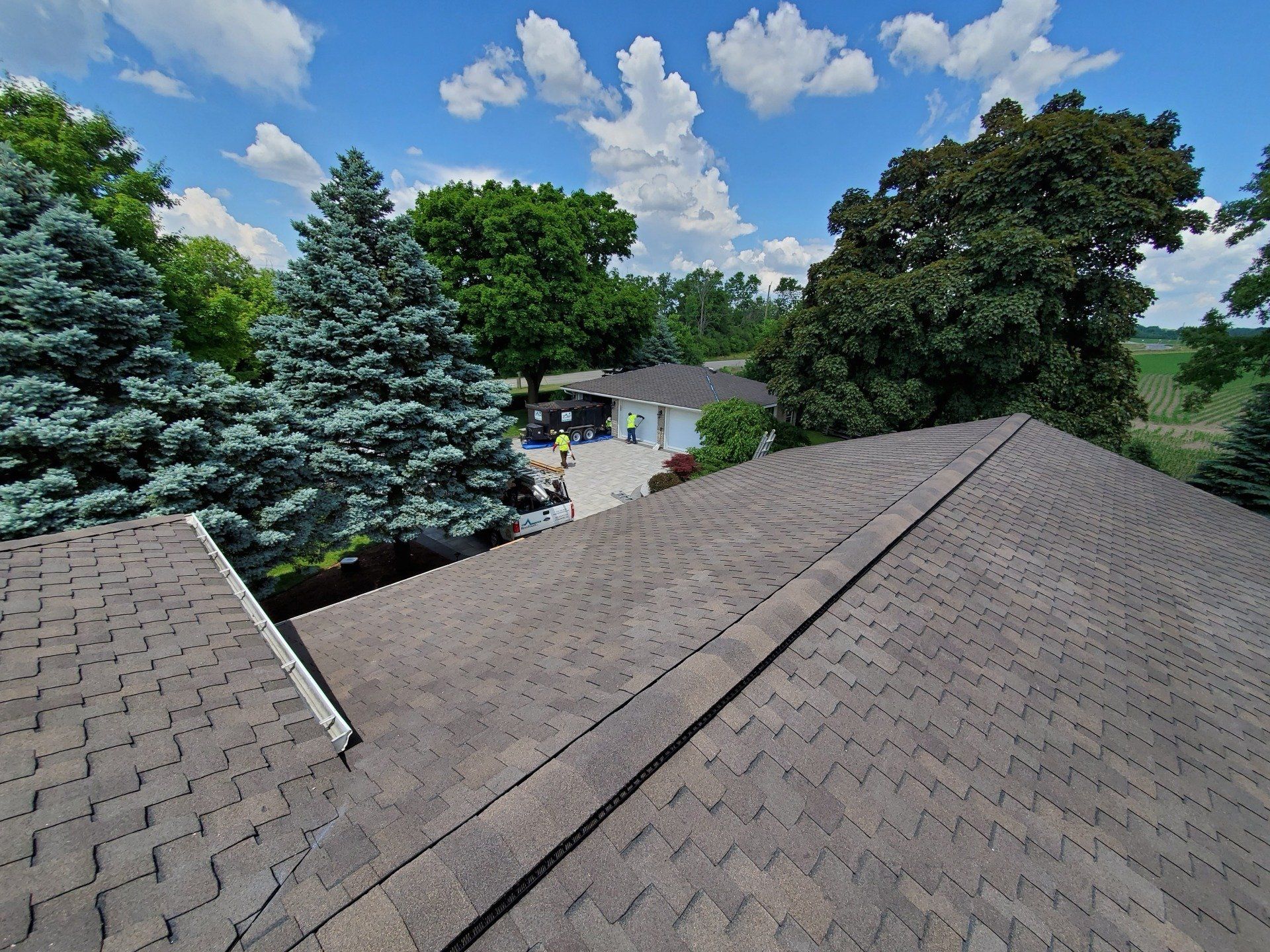 A roof's peak overlooks a residential area with lush greenery, mature trees, a clear blue sky with clouds, and a driveway with vehicles.