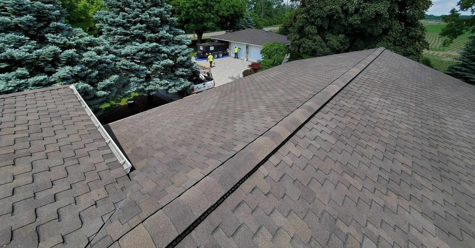 Aerial view of a shingled roof with trees surrounding a property. A person in reflective clothing stands near a vehicle and equipment below.