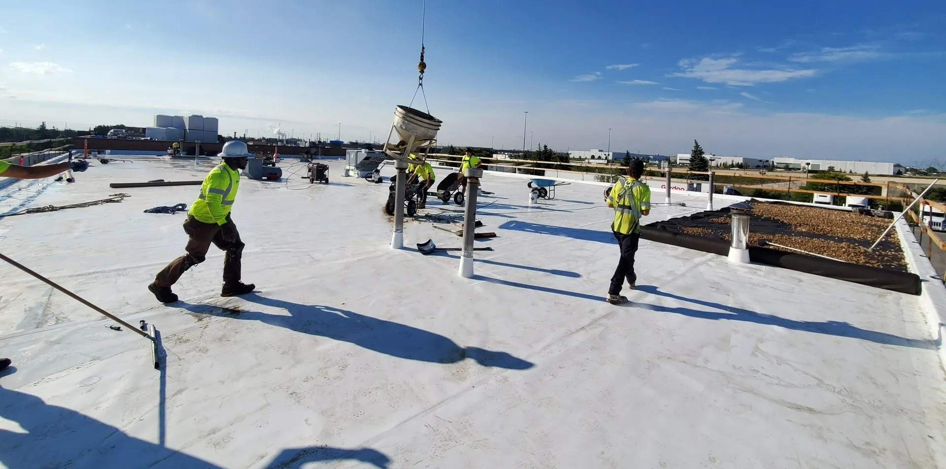 Workers on a commercial roof installing materials, with a crane lifting equipment, under a clear sky, safety gear visible, industrial setting in the background.