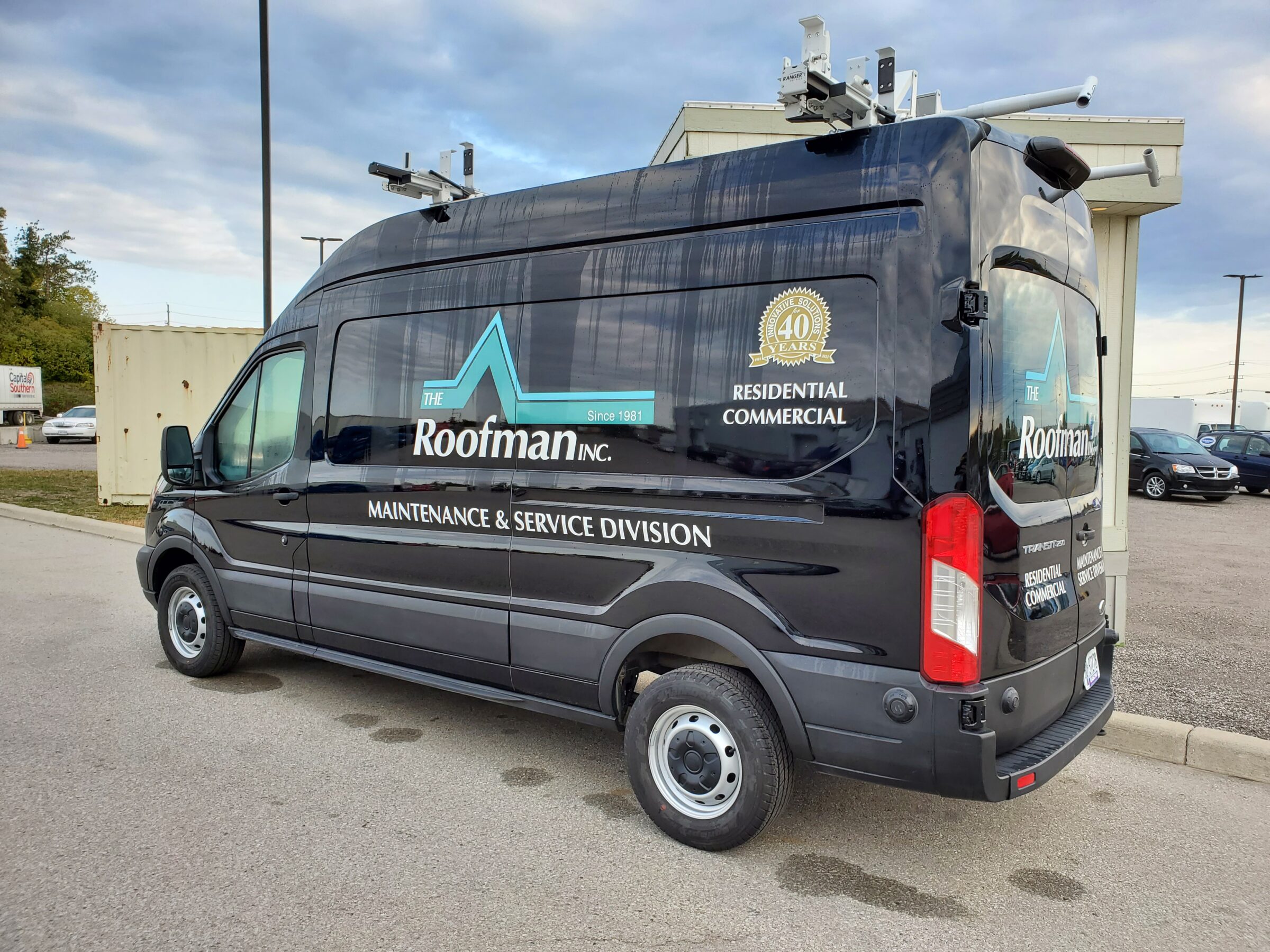 A black commercial van with "The Roofman Inc." branding and logos parked outside, equipped with ladders on top, advertises roofing maintenance and service.