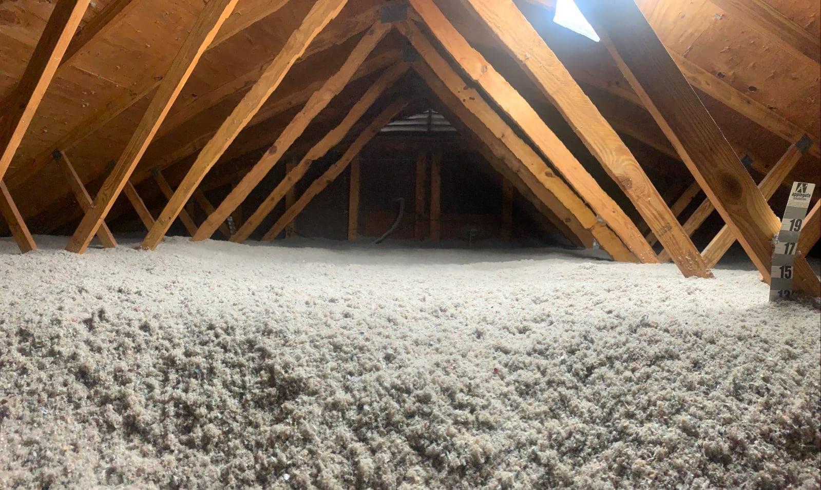 The image shows an attic with wooden beams and rafters. Insulation material covers the floor, and there's a small window, providing some natural light.