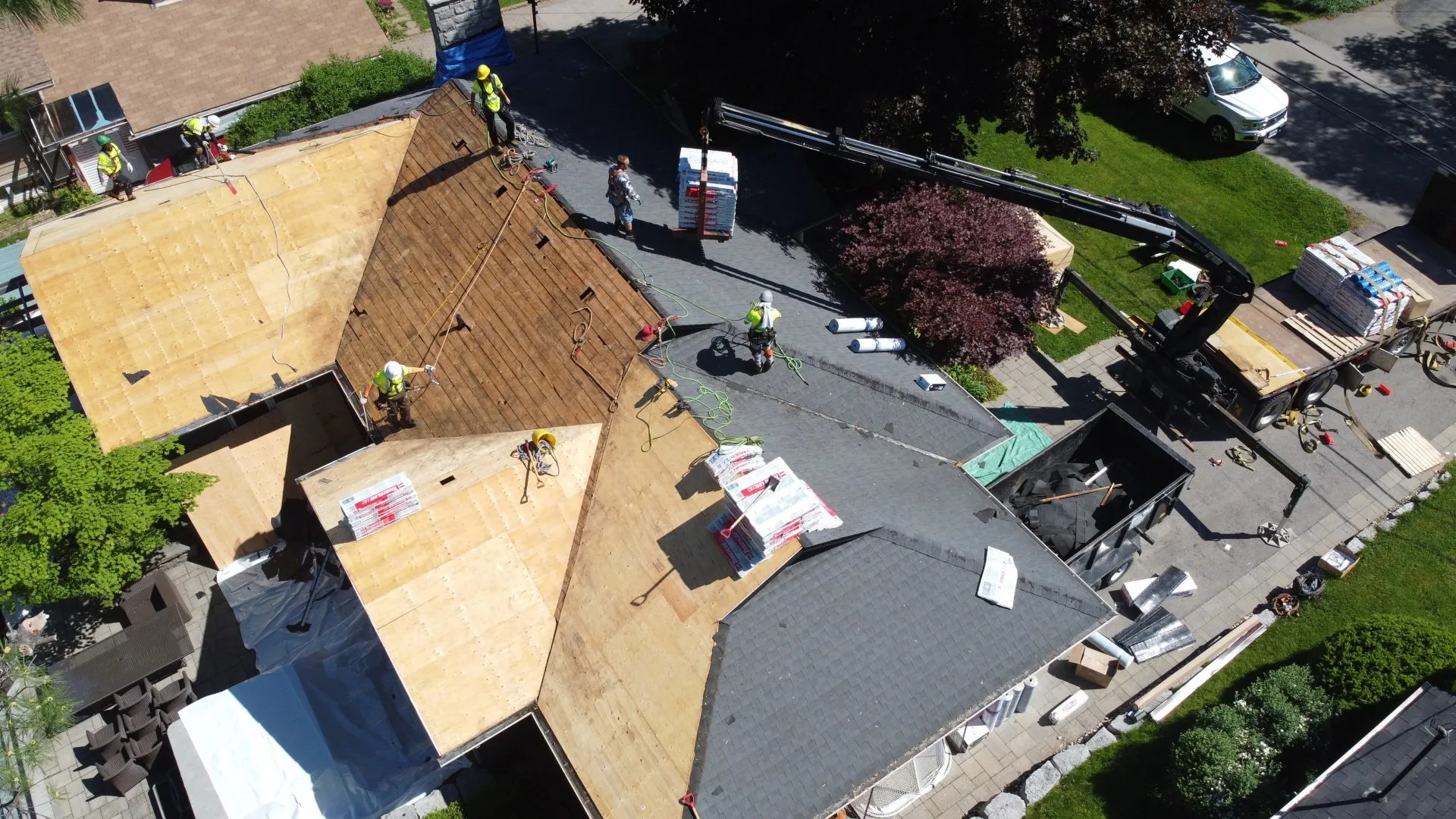 Aerial view of a construction site with multiple people working on a rooftop. Building materials and a crane are visible, with a vehicle parked nearby.