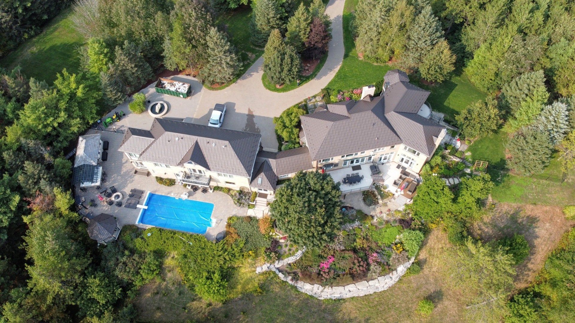 Aerial view of a large multi-level house with a blue pool cover, surrounded by manicured lawns, trees, garden paths, and a driveway with a car.