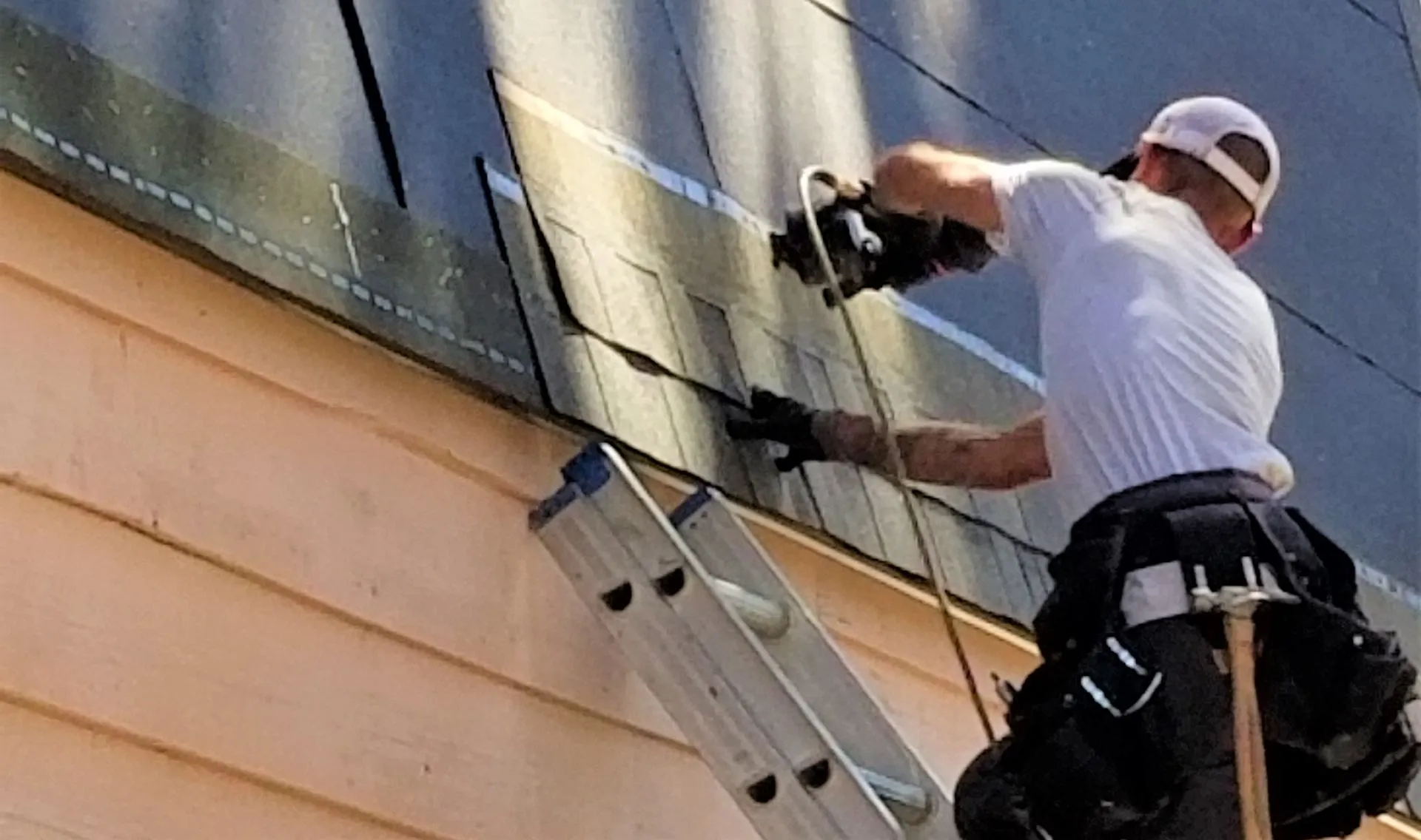 A person is using a nail gun on a roof, standing on a ladder, equipped with a tool belt, wearing a white shirt and a cap.