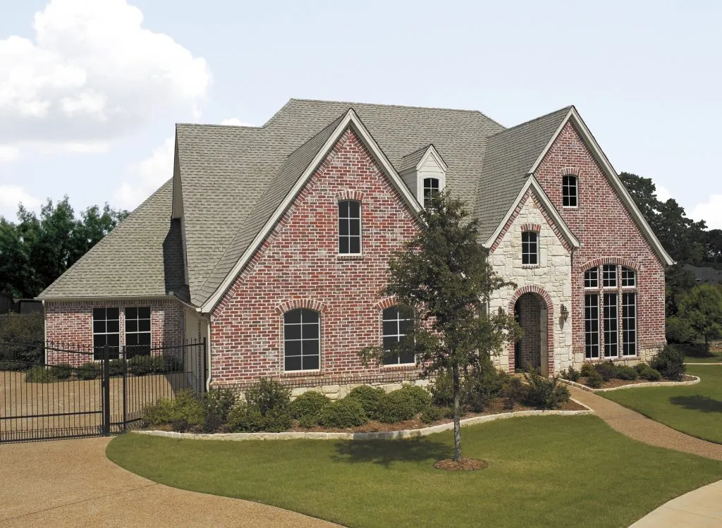 This image shows a large two-story brick house with arched windows, gabled roofs, an attached garage, and a landscaped yard with a small tree.