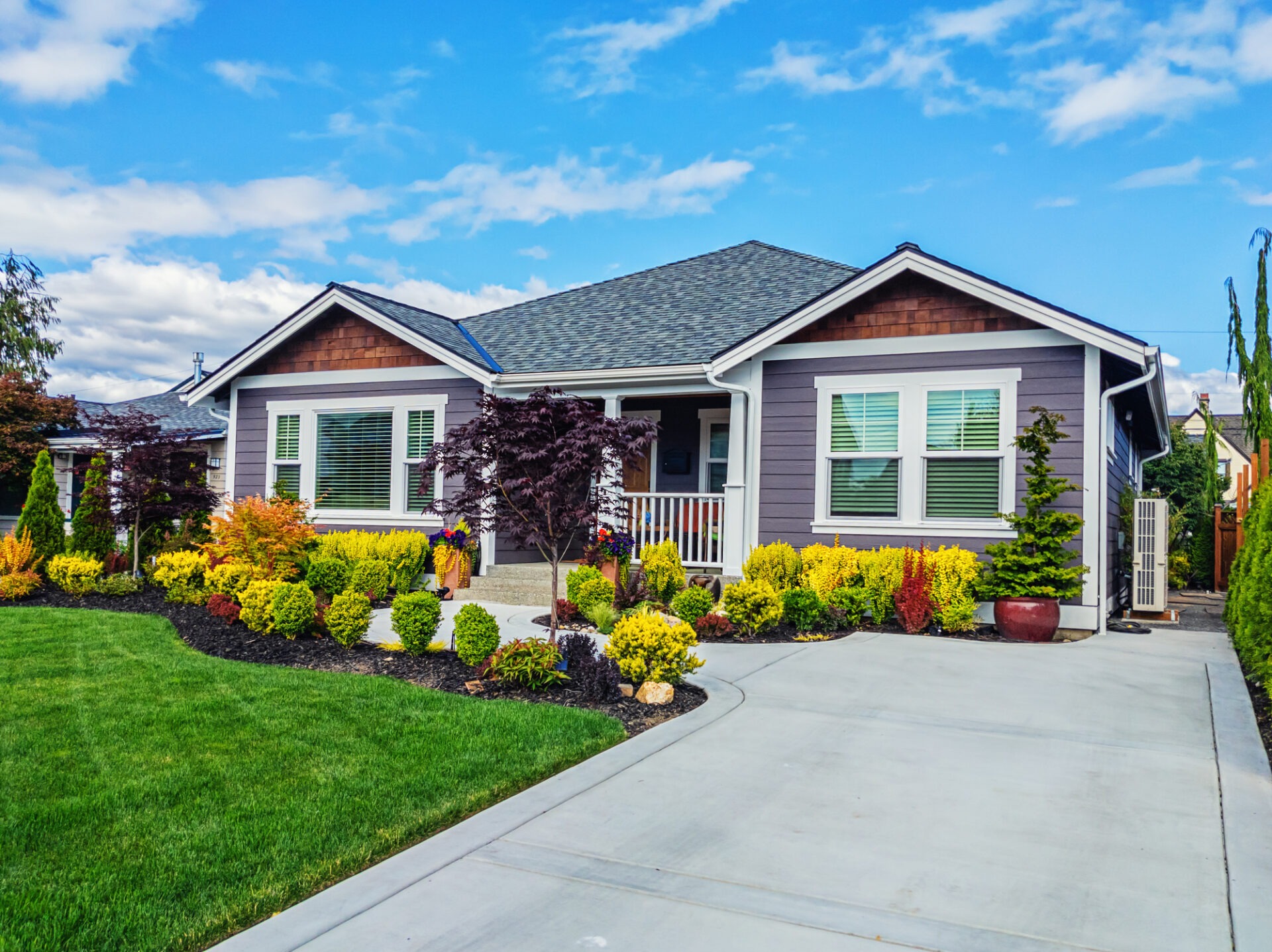 This image depicts a single-story suburban home with a neatly landscaped yard, colorful shrubs, green lawn, a driveway, and a clear blue sky.