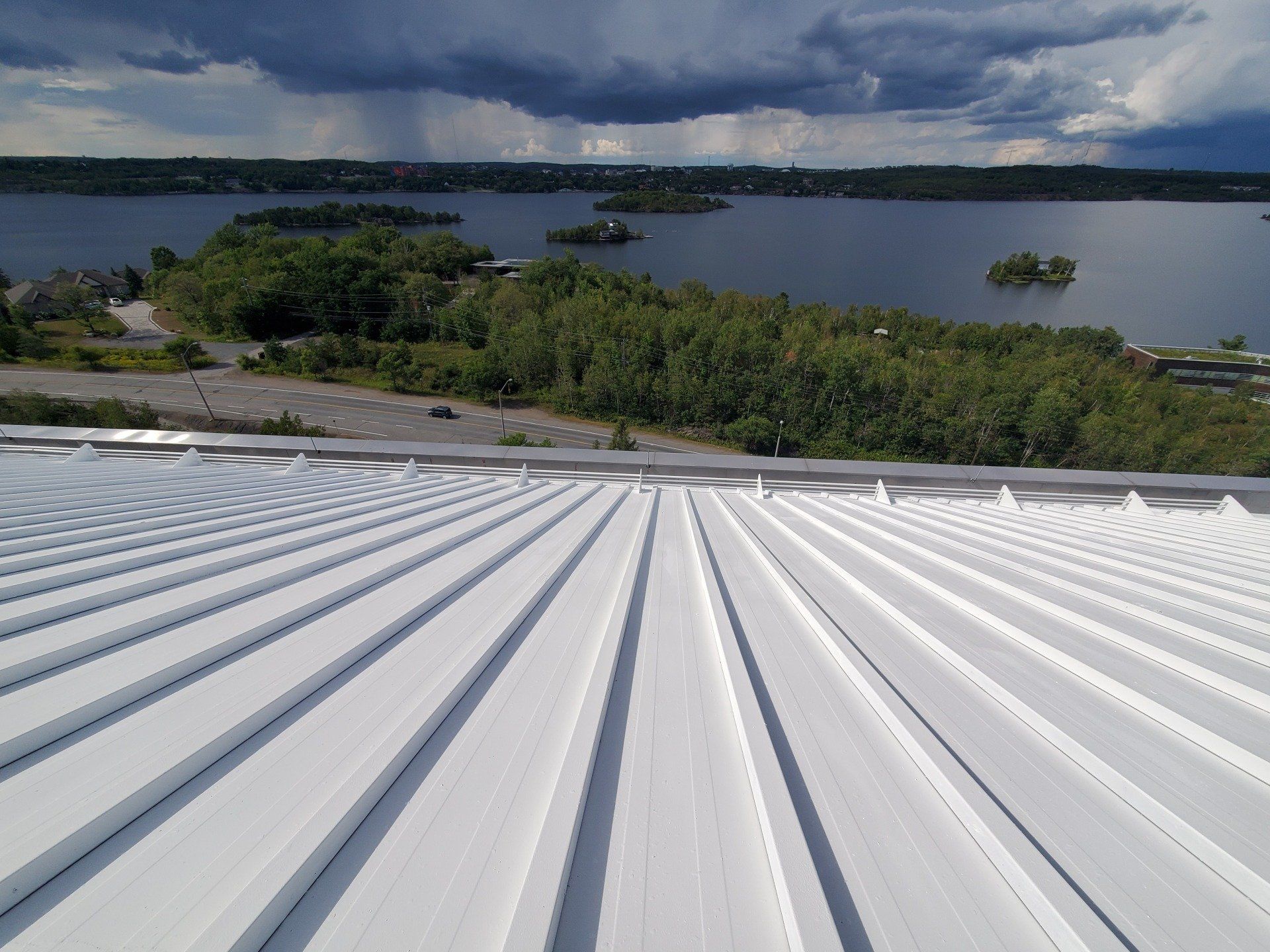 An aerial view over a metal roof with ridges, overlooking trees, a road, and a lake under a cloudy sky, with a rain shower in the distance.