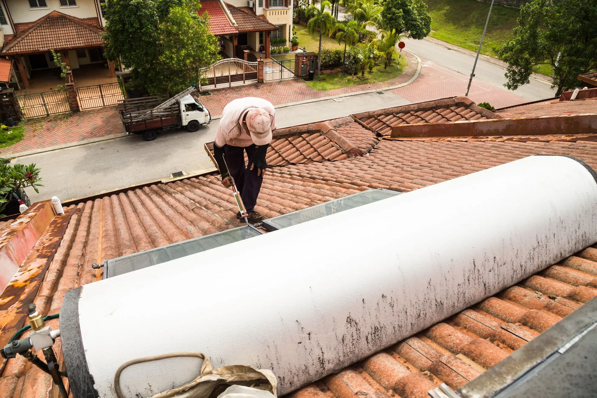 A person is working on a tile roof, installing insulation material, wearing a pink cap and gloves, with suburban houses and a truck below.