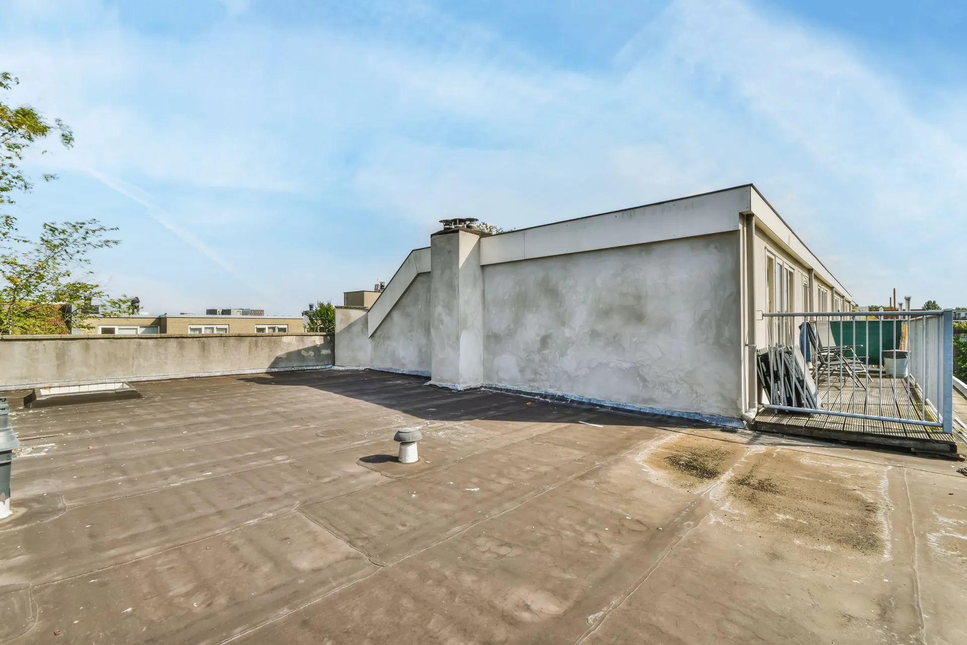 Empty flat rooftop with railing, chimney, vents, and clear blue sky. Building structure visible with a metal stairway leading down. No people present.