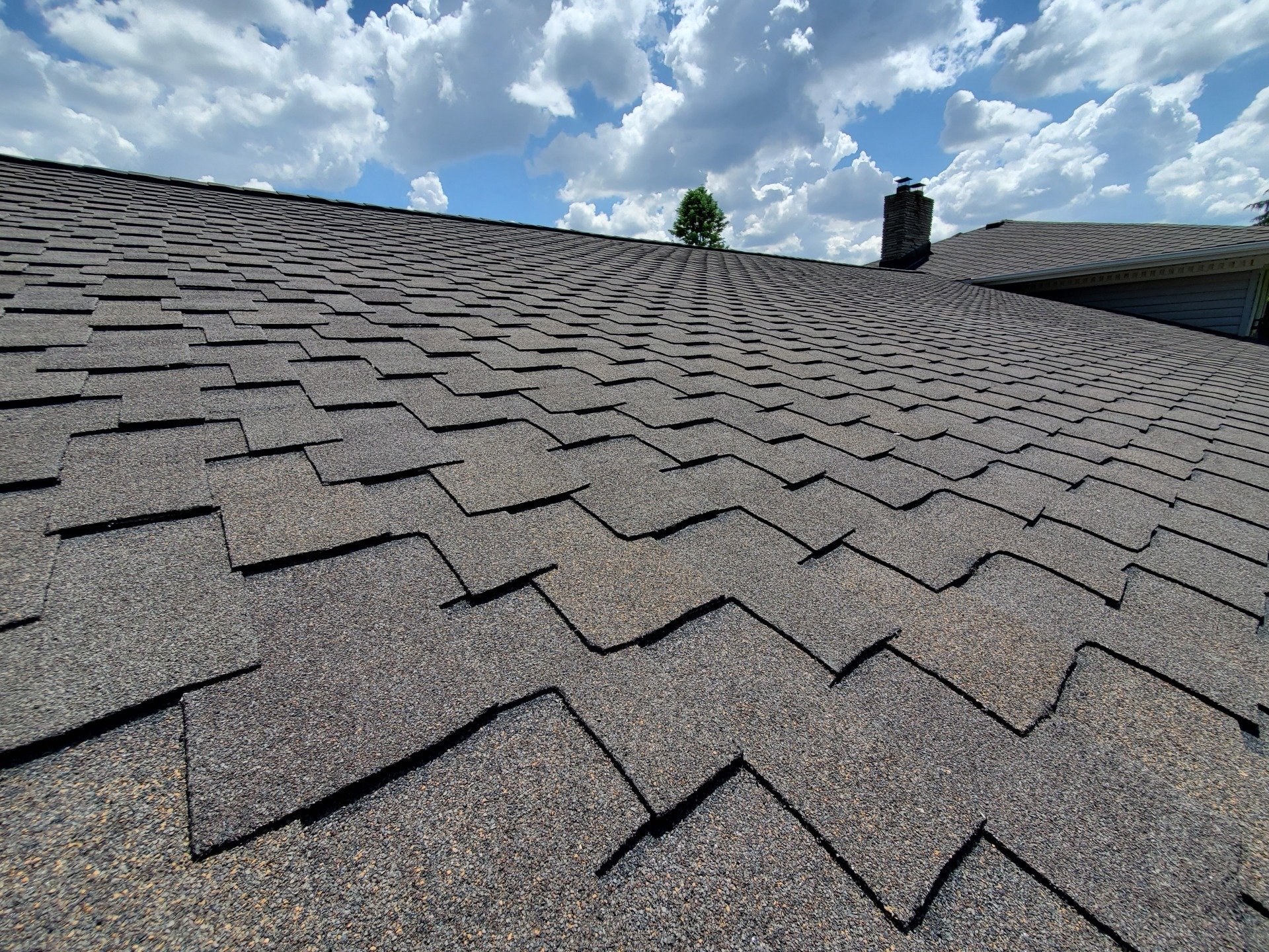 The image shows a close-up of an asphalt shingle roof with a chimney, clear blue sky, white clouds, and a hint of a tree visible.