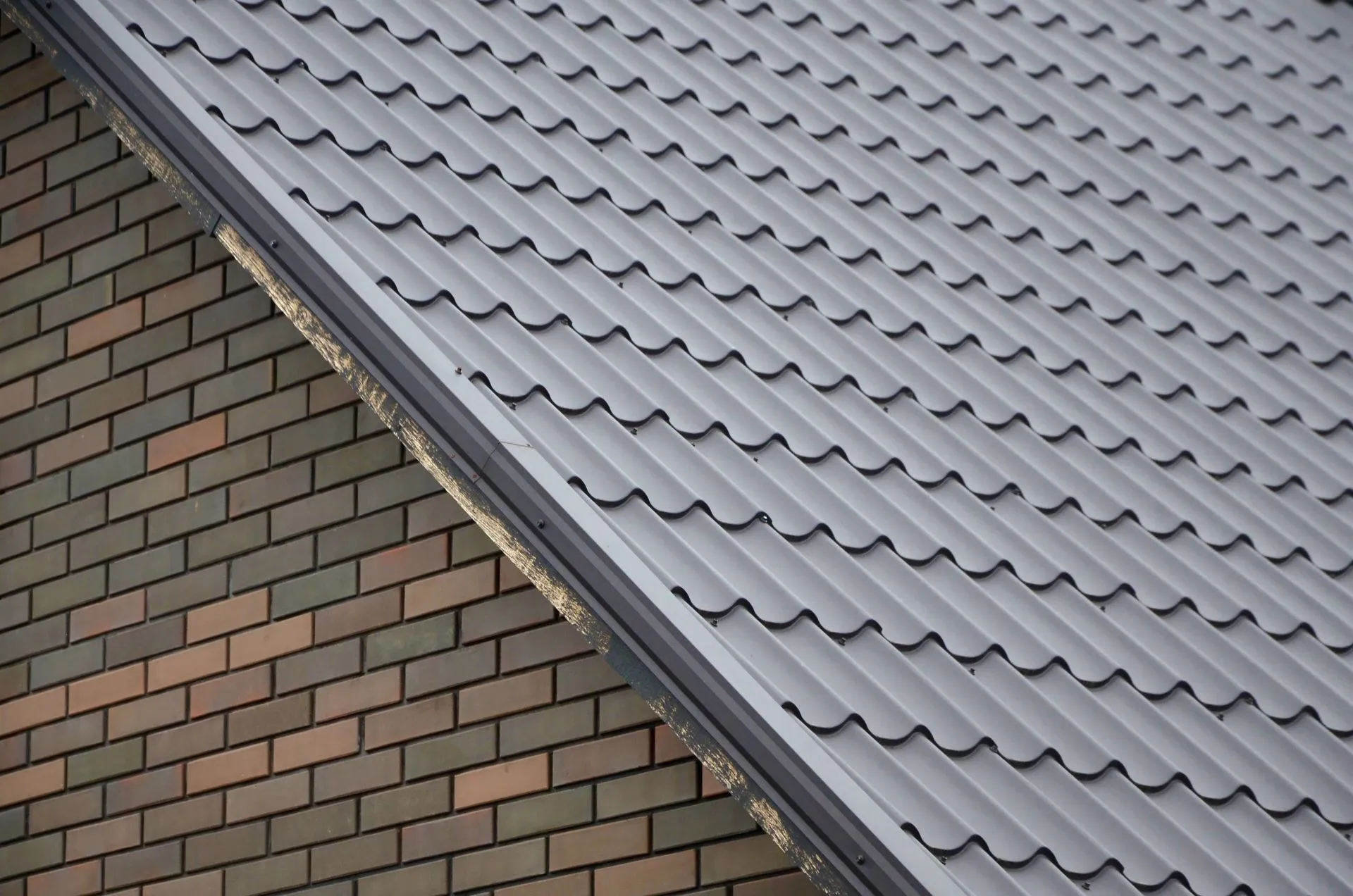 This image shows a close-up of a building's corner, where a gray corrugated metal roof meets a brick wall with varying shades of brown bricks.