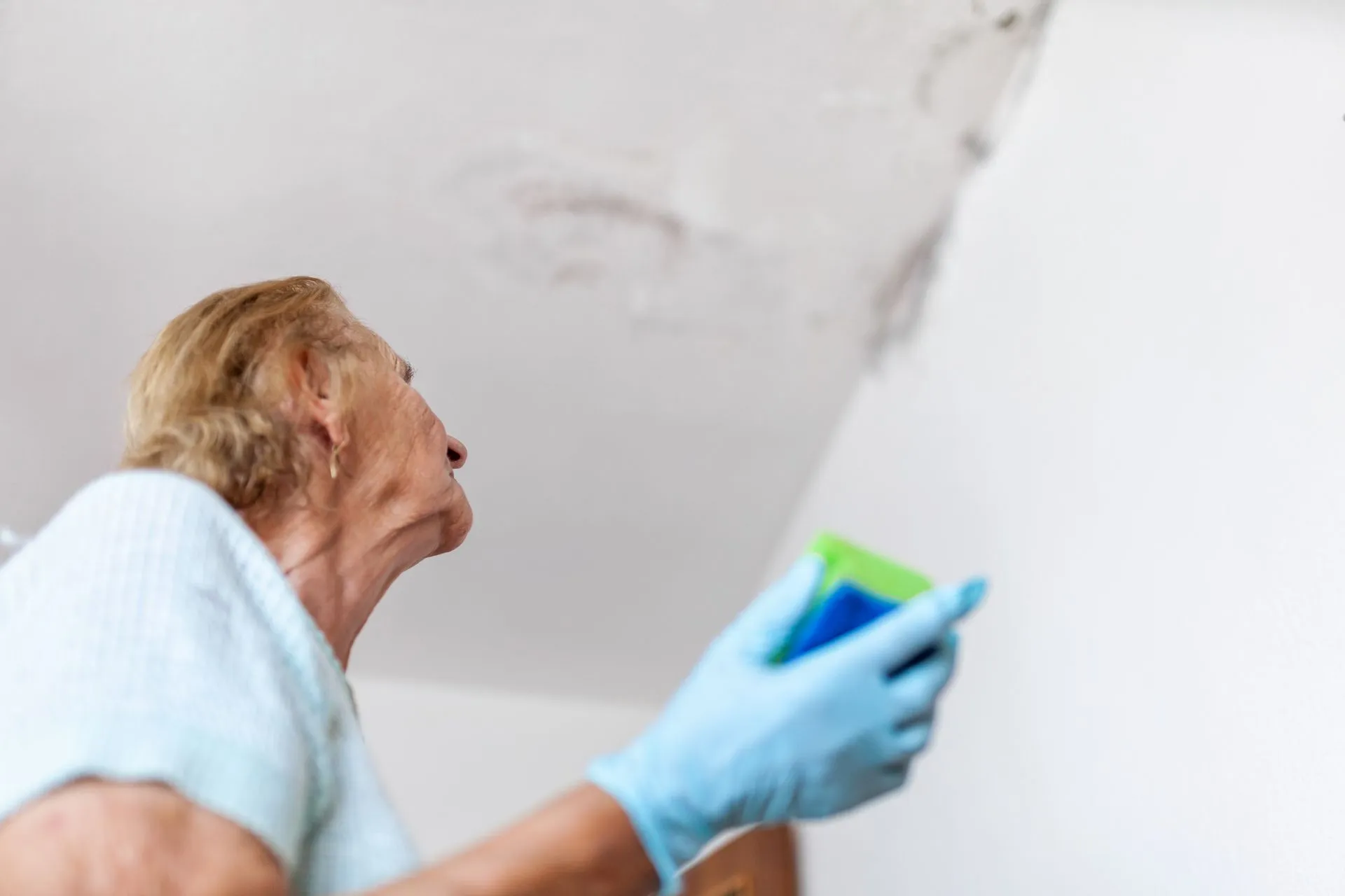 A person is inspecting a ceiling with water damage, holding a sponge, wearing blue gloves, likely preparing to clean or repair the area.