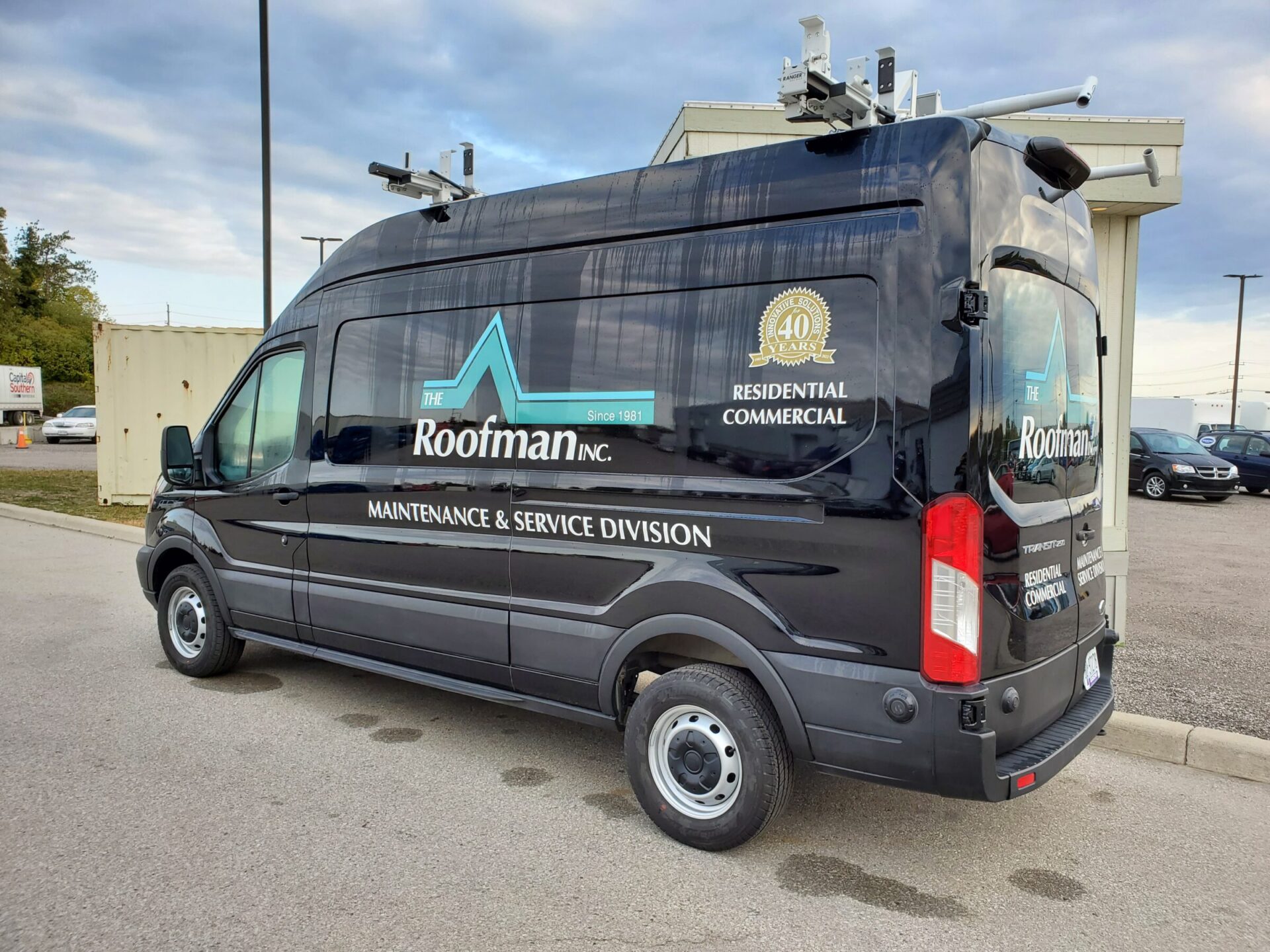 A commercial black van with business branding for "The Roofman Inc." equipped with ladders and maintenance gear, parked in a lot under a clear sky.