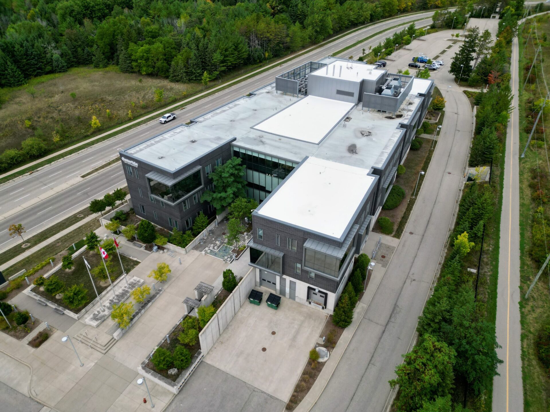 Aerial view of a modern office complex with flat roofs next to a highway, surrounded by greenery, under overcast skies, with a few vehicles parked.