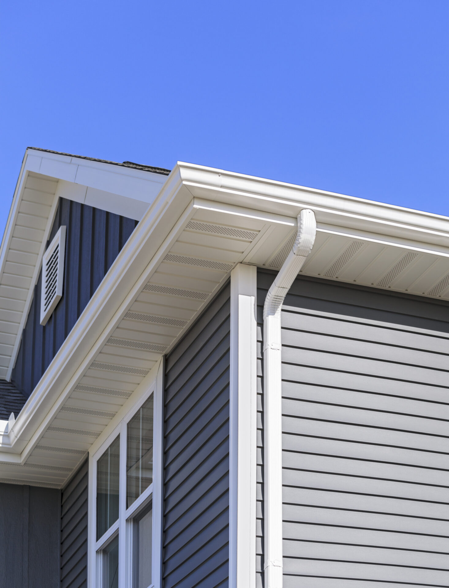 A section of a house showing vinyl siding, two windows, and a gutter downspout against a clear blue sky. Elements of modern suburban residential architecture are visible.