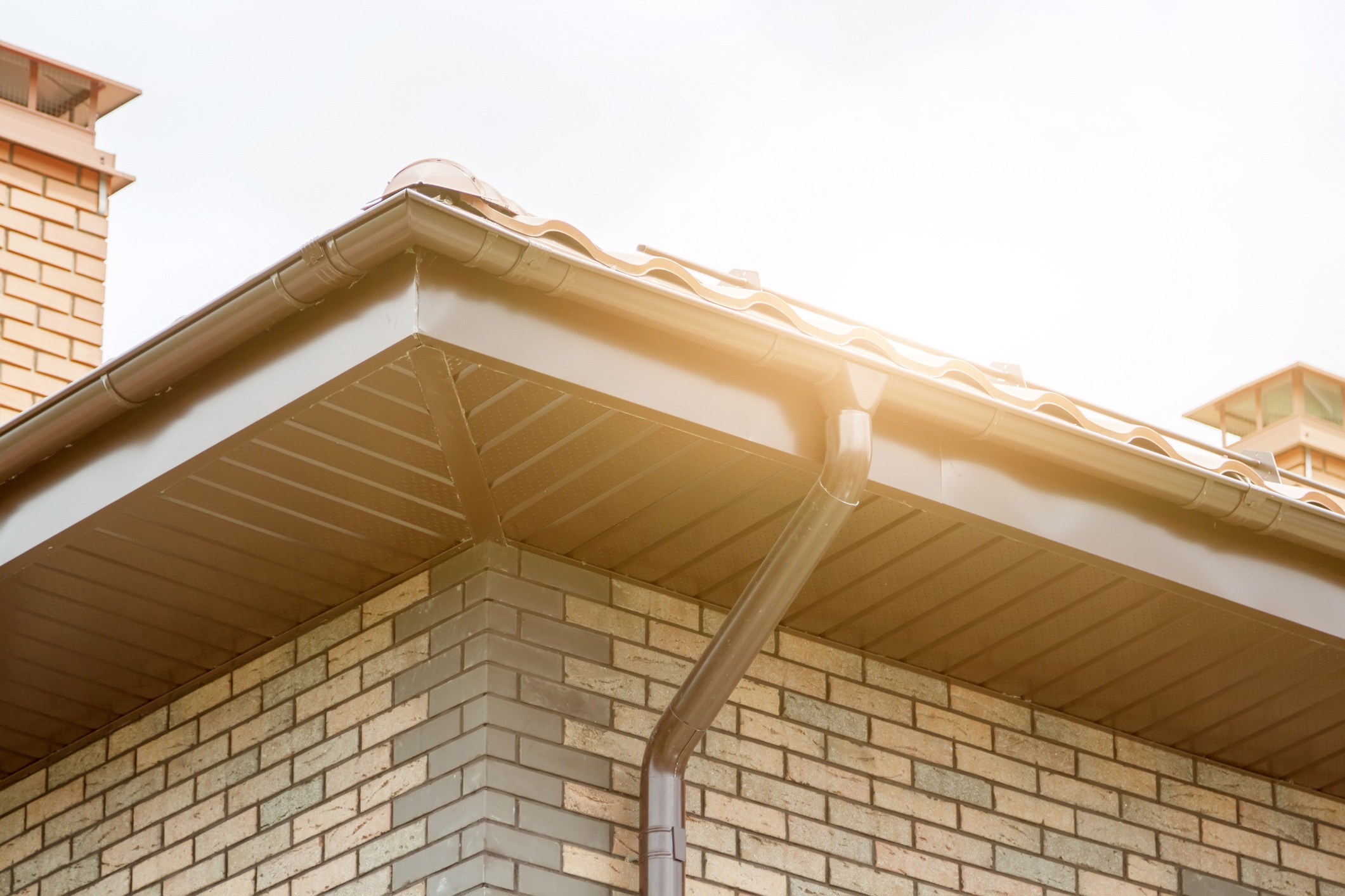 The image shows a close-up of a house's roof corner with a gutter and downspout against a brick wall, bathed in warm sunlight.